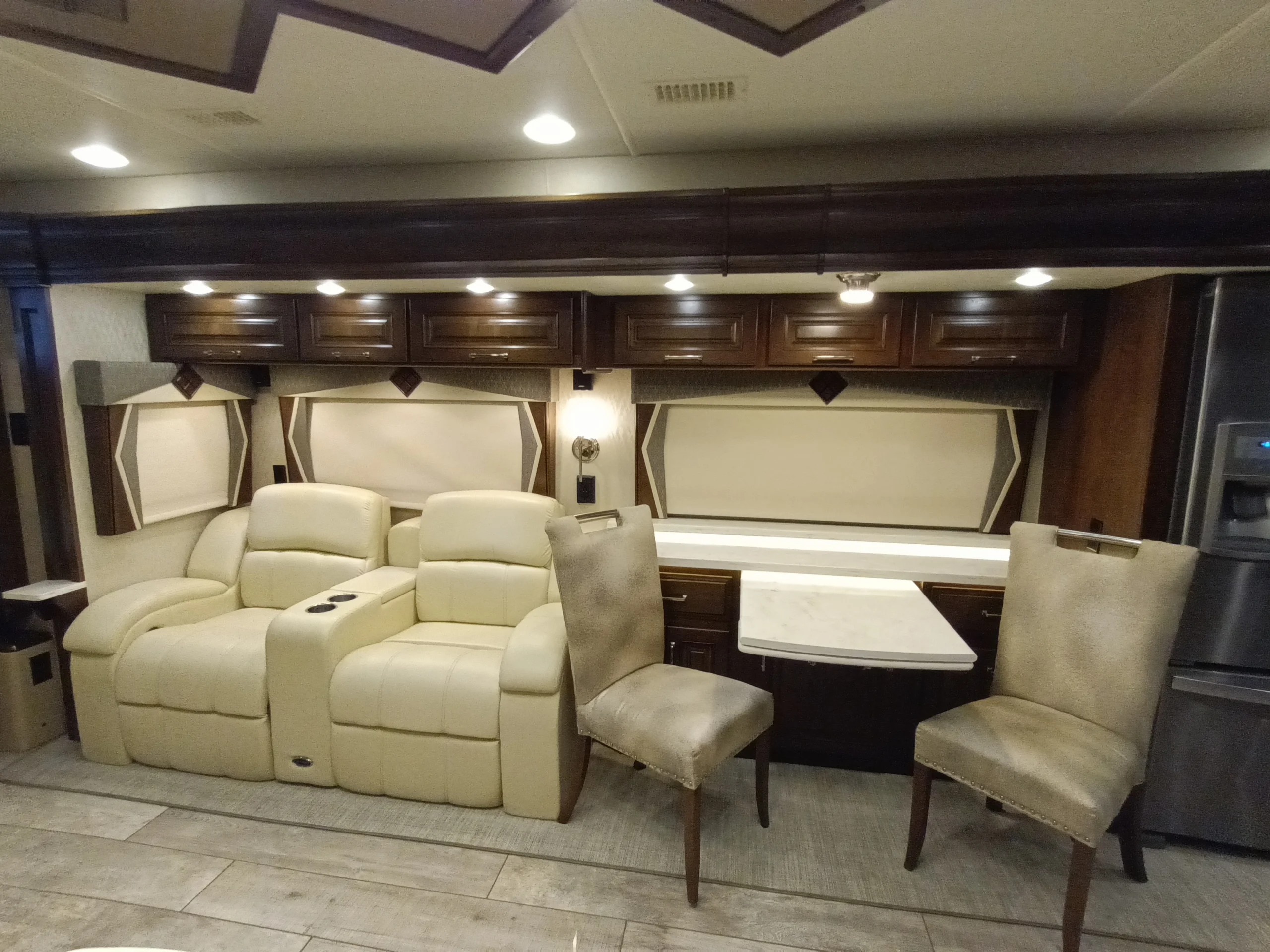 IMG_20260604_122329_595 Luxury Motorcoach interior with beige recliners, two dining chairs, wood cabinets, and a compact table.