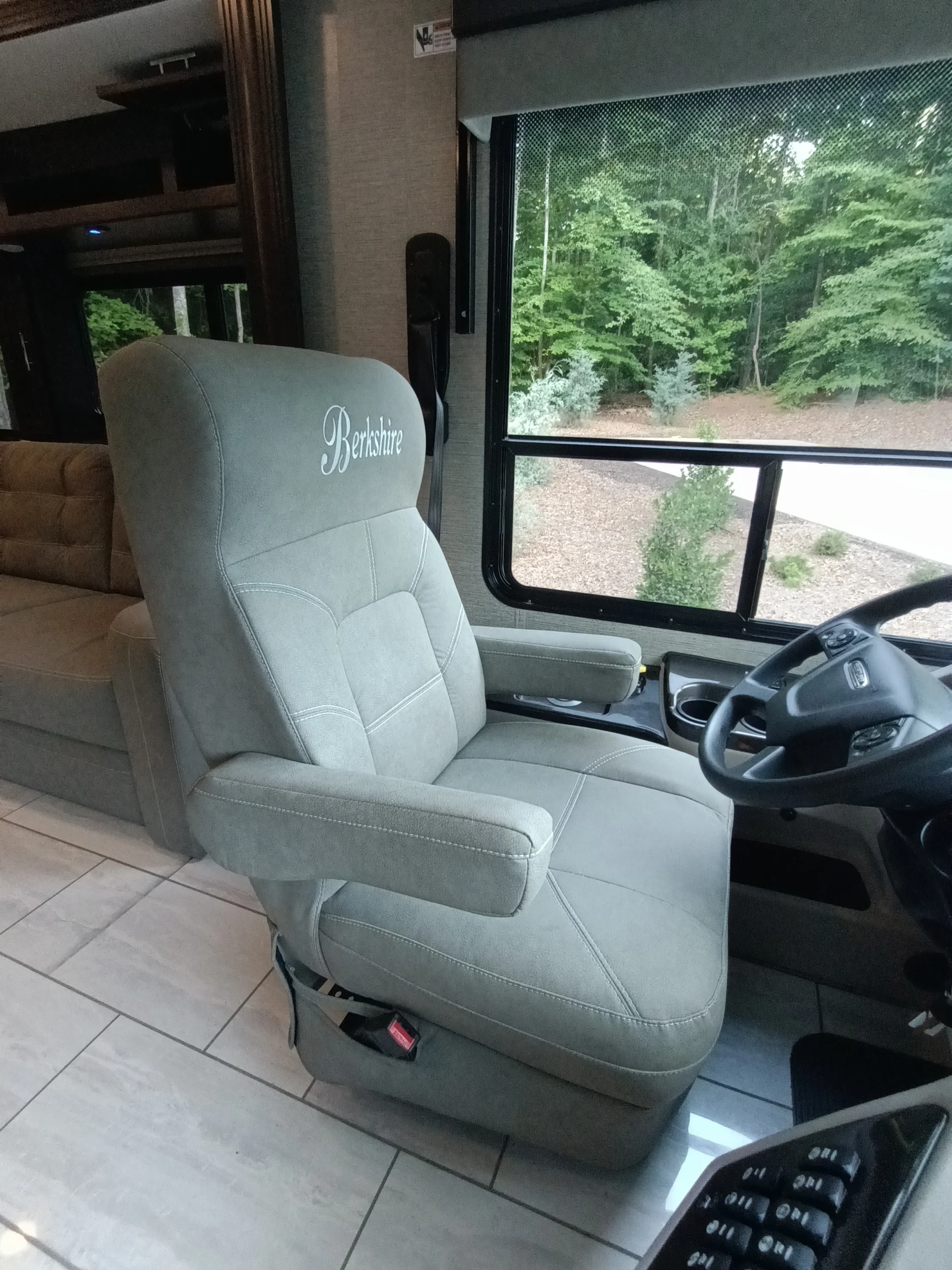 IMG_20260611_190554_911 Motorcoach driver's seat labeled Berkshire, steering wheel nearby, large window showing forest, interior includes couch and tiled floor.