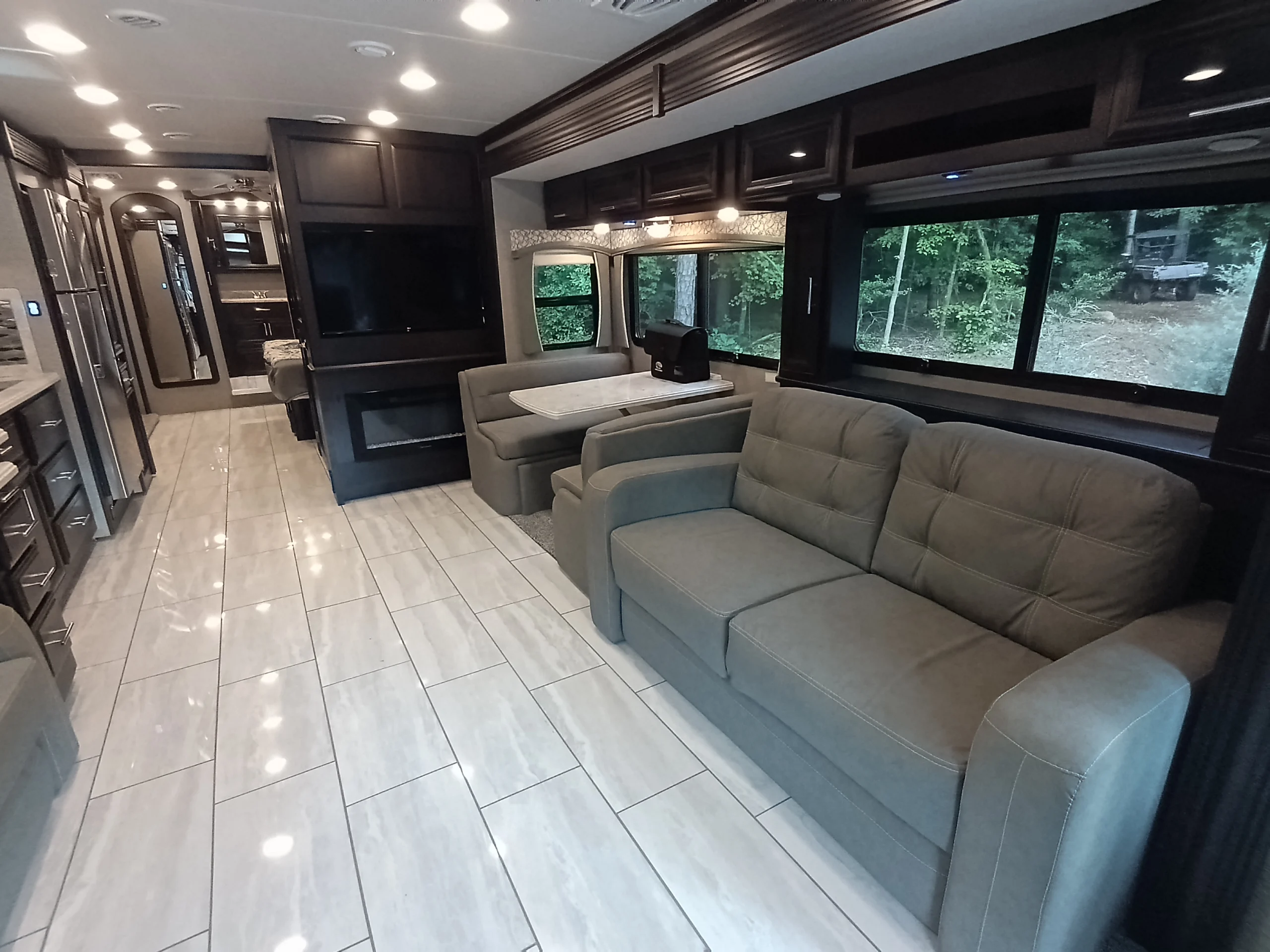 IMG_20260611_190638_053 Luxurious Motorcoach interior featuring a tile floor, plush sofa, dining area, flat-screen TV, and large windows with a forest view.