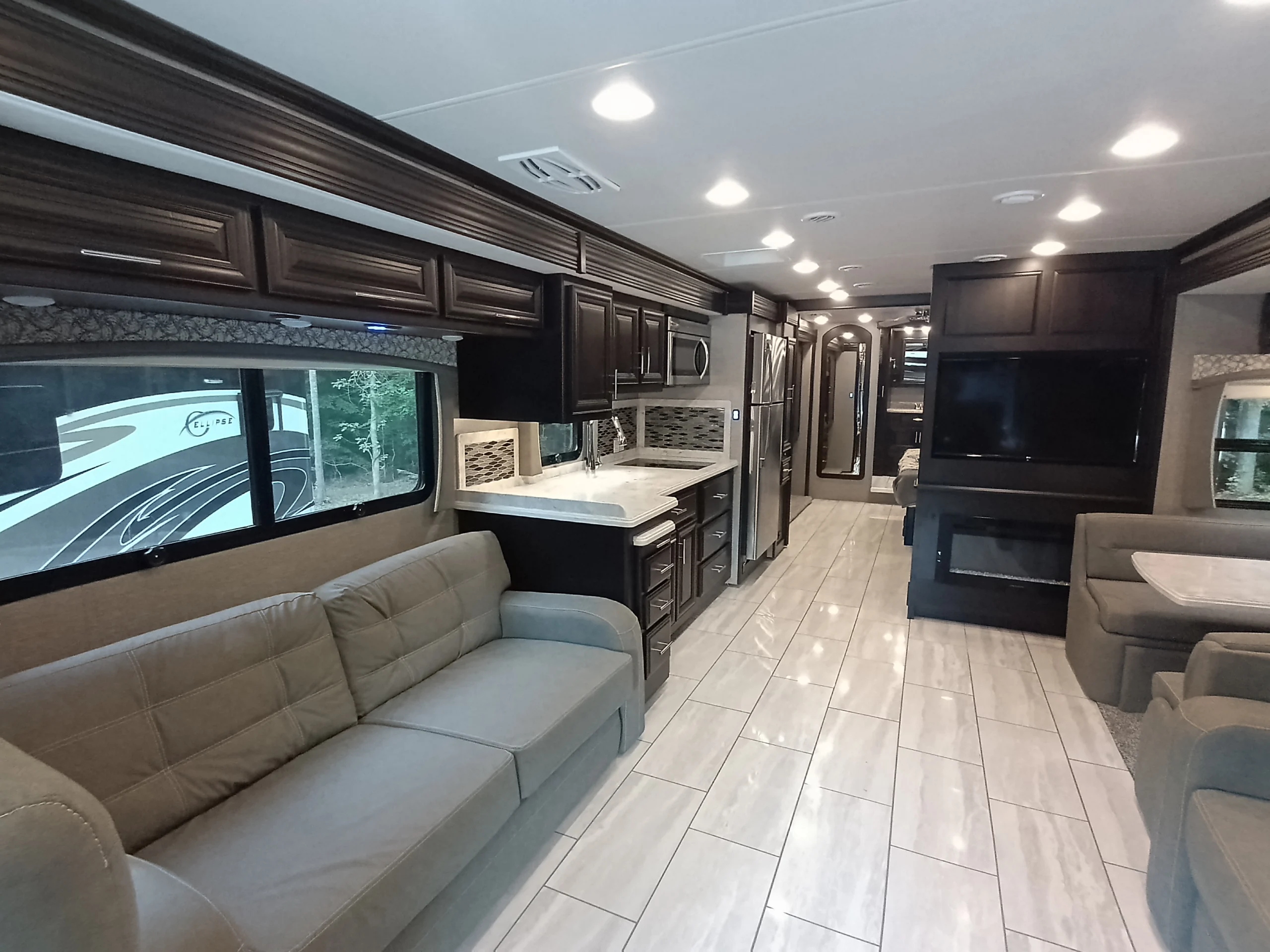 IMG_20260611_190715_437 Spacious Motorcoach interior with modern kitchen, dark cabinets, large fridge, cozy seating, entertainment system, and tiled flooring.