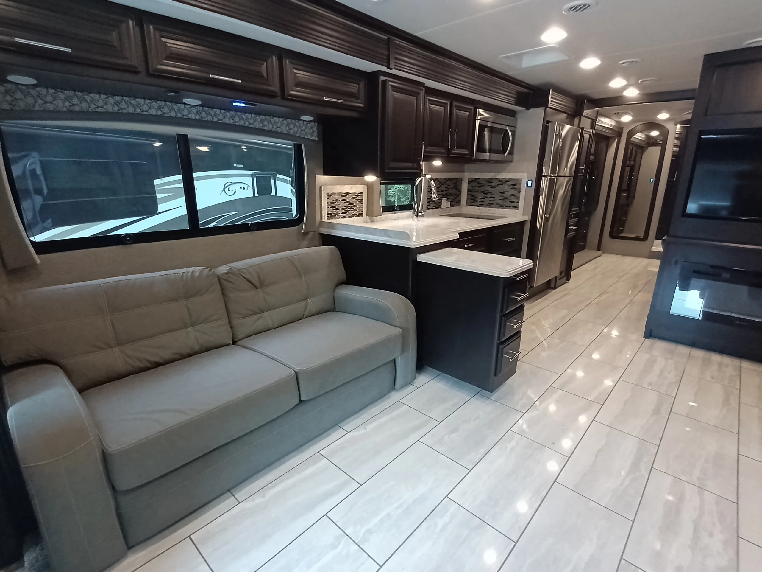 IMG_20260611_190734_916 Luxurious Motorcoach and RVs interior with a leather sofa, modern kitchen, stainless steel appliances, and glossy tiled flooring.