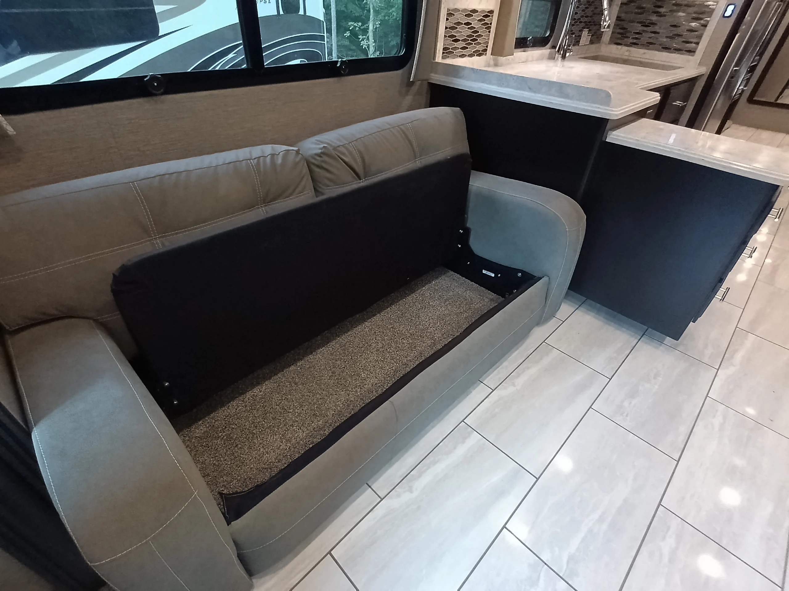 IMG_20260611_190814_113 Gray RV sofa with removable cushion, revealing storage space. Adjacent kitchen area with light tiled flooring. Motorcoach and RVs interior.