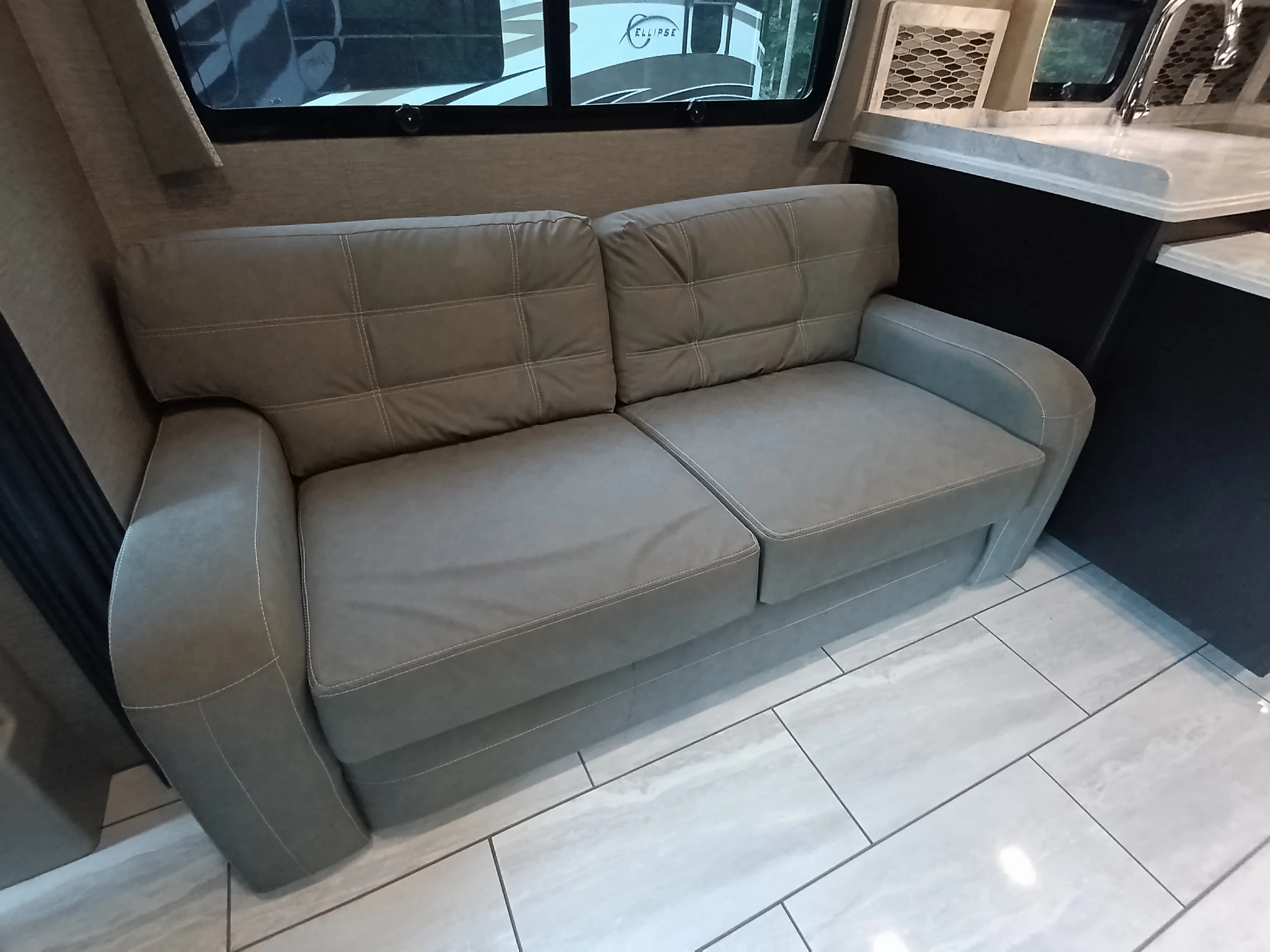 IMG_20260611_190841_820 Gray cushioned sofa inside a Motorcoach and Rvs, next to a kitchenette with a tiled floor.