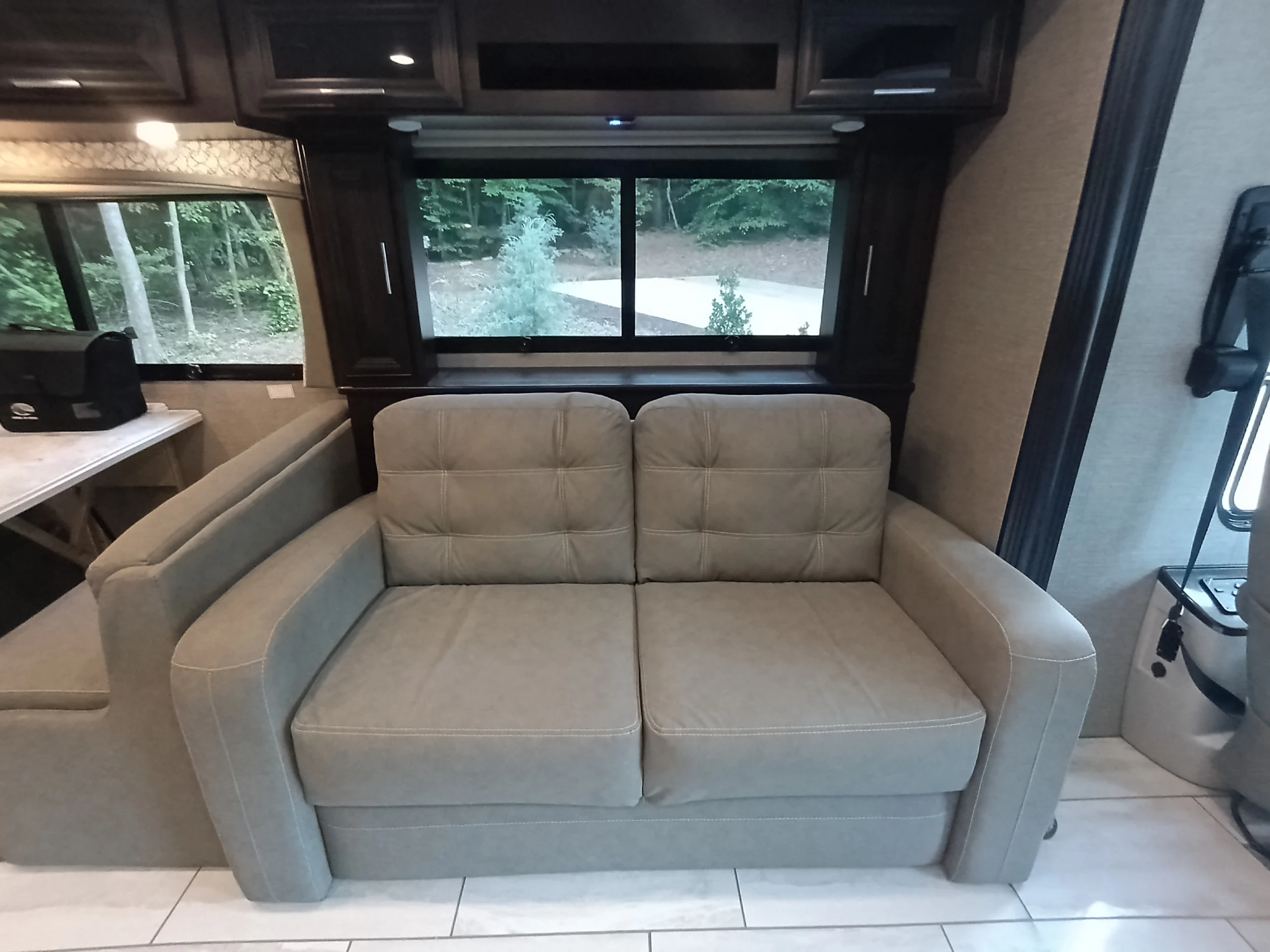IMG_20260611_190849_096 Beige couch in a Motorcoach and RVs interior, situated by a window with an outdoor view, adjacent to a dining area with a black bag.