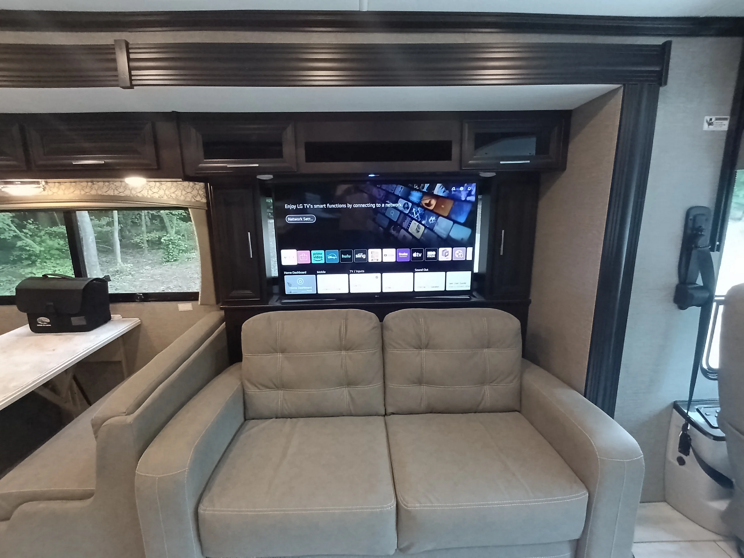 IMG_20260611_190946_193 Luxurious Motorcoach interior featuring a cozy couch, wall-mounted smart TV, dining area, and large windows with scenic outdoor view.