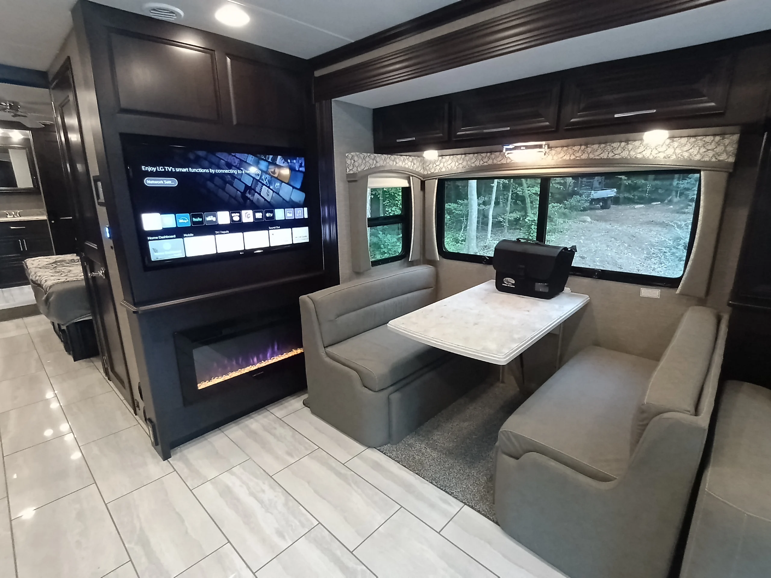 IMG_20260611_191017_351 Motorcoach interior with TV, electric fireplace, dining area, and large windows showcasing wooded view.