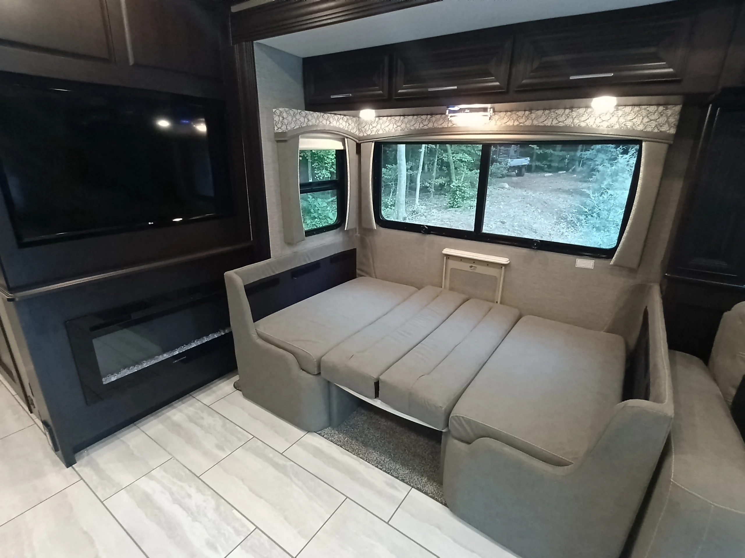 IMG_20260611_191101_937 Spacious interior of a motorcoach, featuring a fold-out bed, large window with a forest view, wall-mounted TV, and modern fireplace.