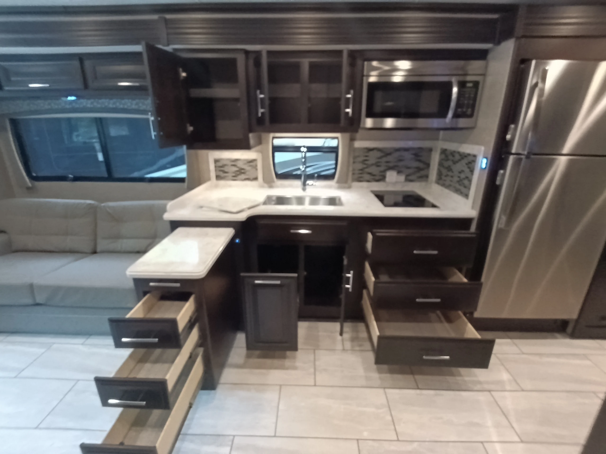 IMG_20260611_191231_399 Spacious Motorcoach kitchen with open cabinets, drawers, stainless steel appliances, and a sofa by a large window.