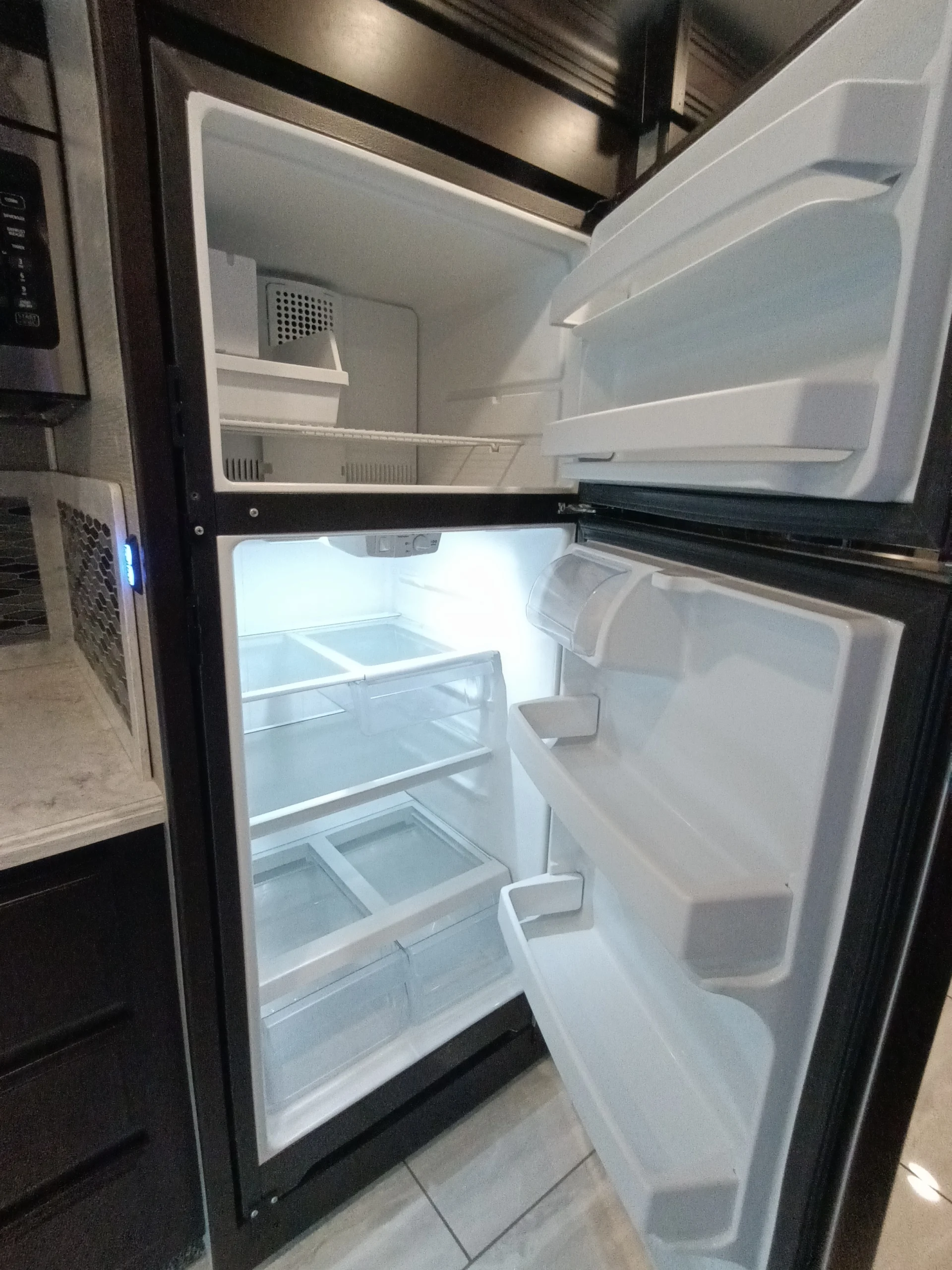 IMG_20260611_191327_042 Open RV refrigerator in a motorcoach, showing empty freezer and fridge sections with shelves and door compartments.
