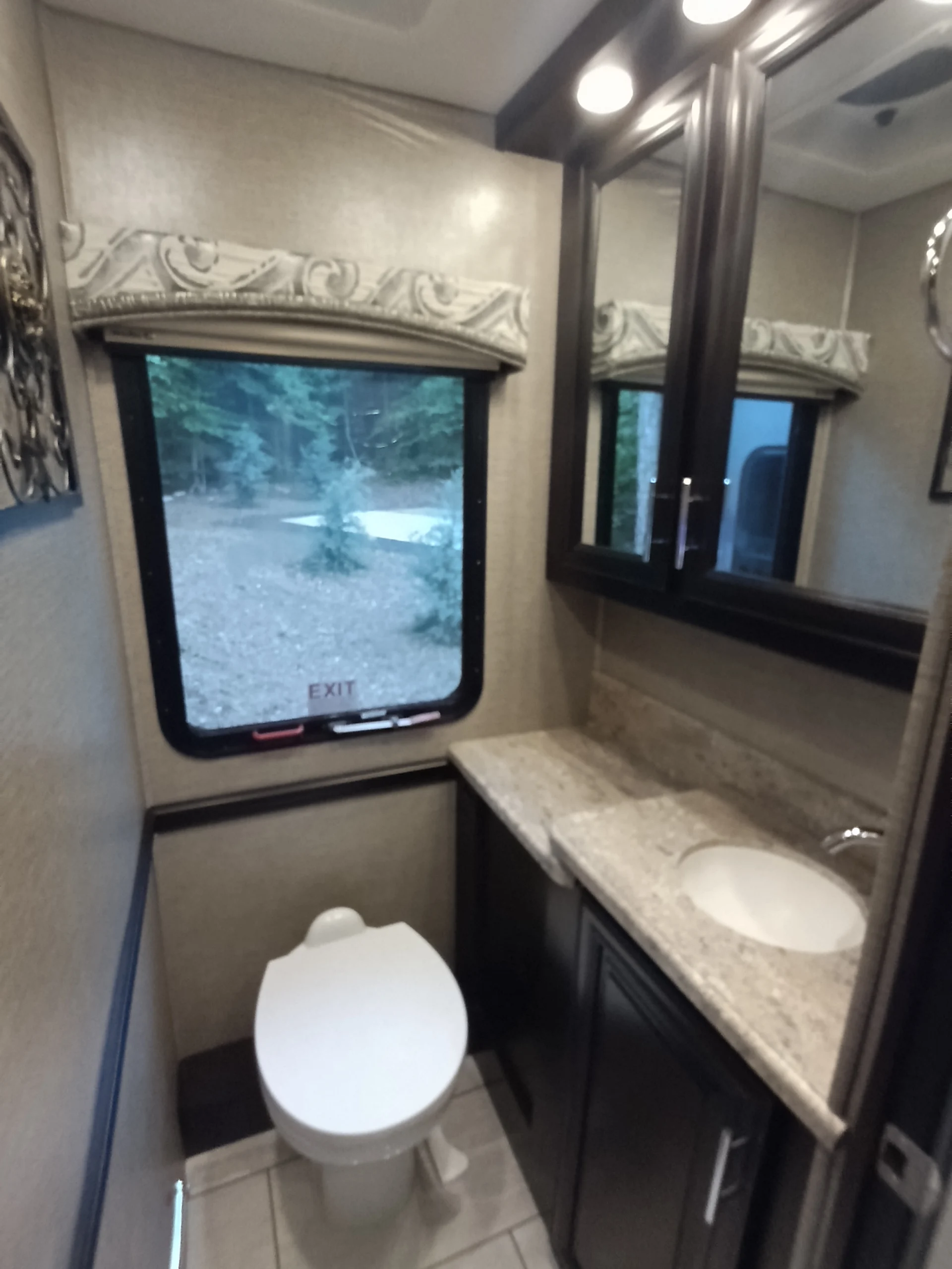 IMG_20260611_191540_679 RV bathroom with modern fixtures; includes a toilet, marble countertop, sink, mirror, and window with a forest view.