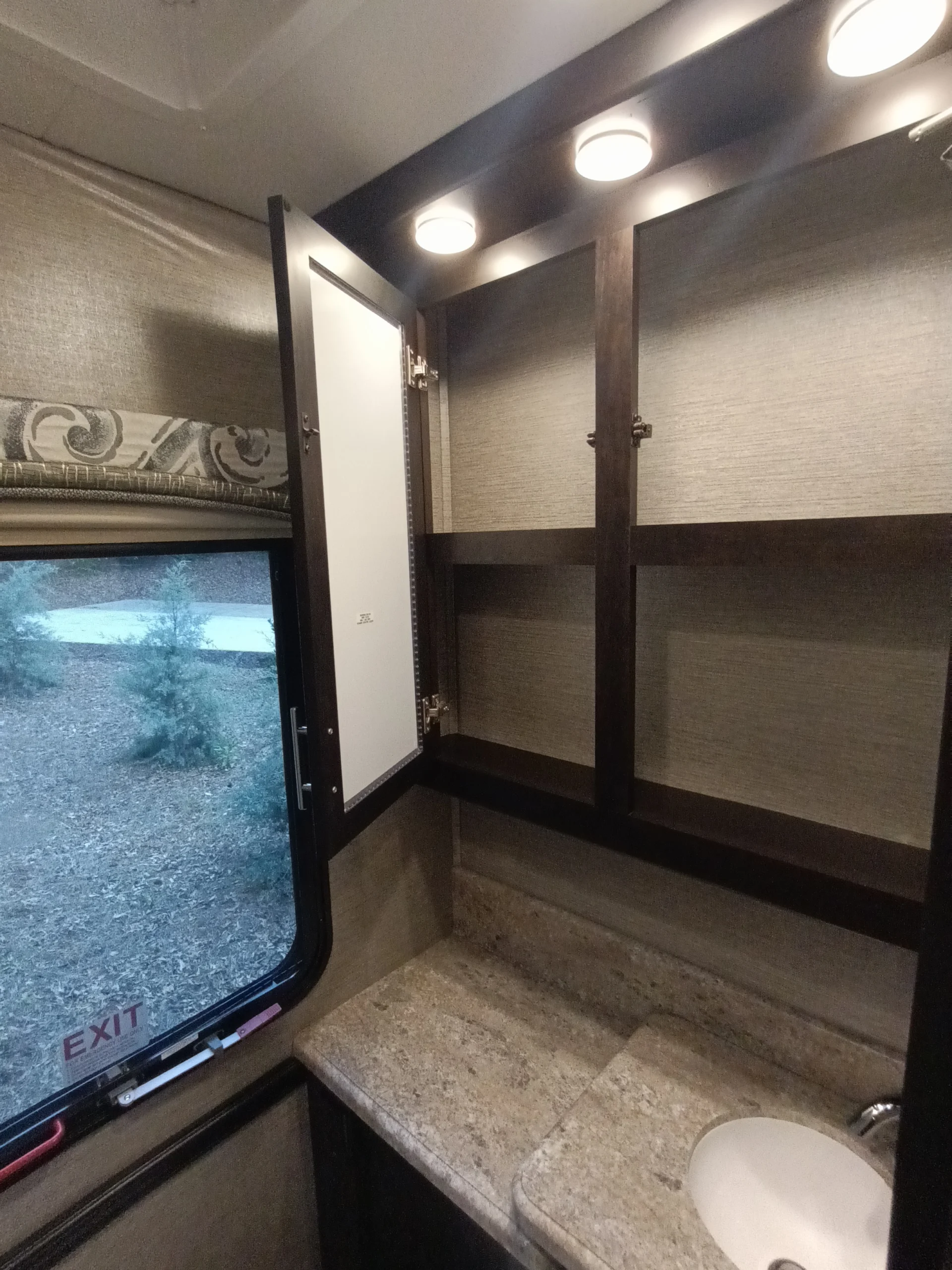 IMG_20260611_191553_658 RV bathroom features open cabinet, marble countertop with sink, window with exit sign, and overhead lights, typical of motorcoaches.