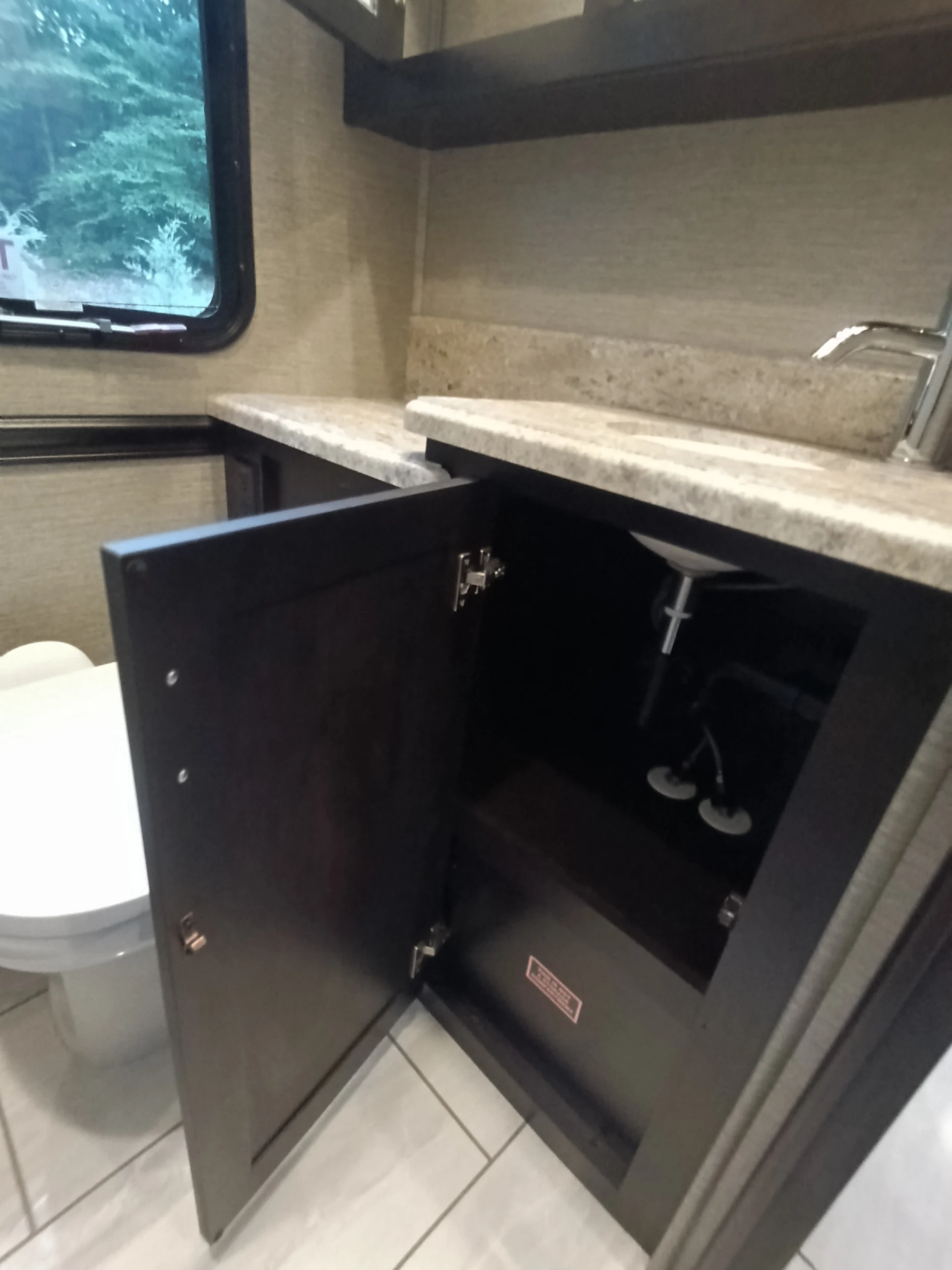 IMG_20260611_191558_439 Motorcoach bathroom with open wood cabinet, white toilet, marble countertop, stainless faucet, and window view.
