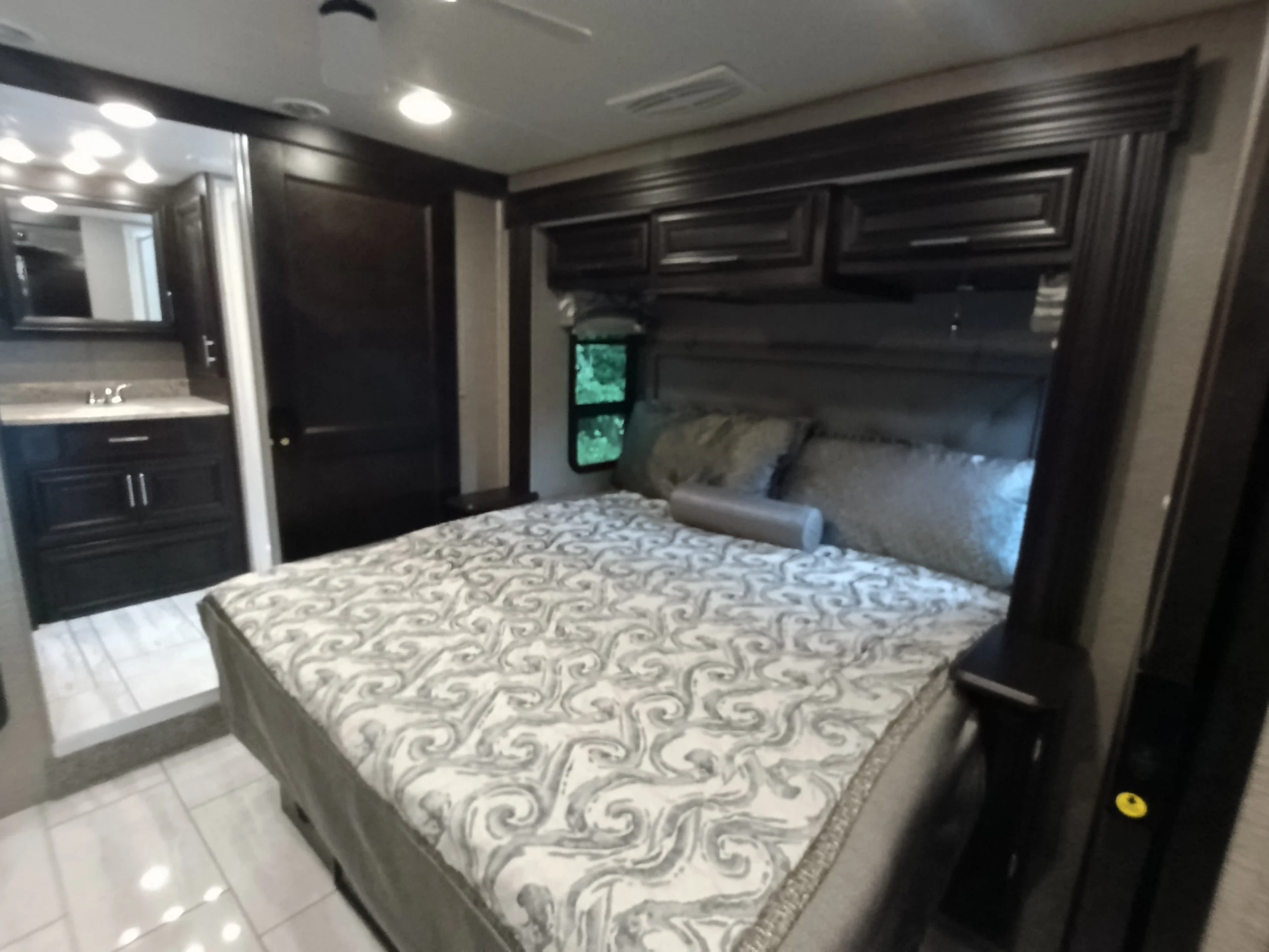 IMG_20260611_191721_067 Luxury RV bedroom with a patterned bedspread, overhead cabinets, and adjacent bathroom with sink and mirror. Motorcoach and RVs.