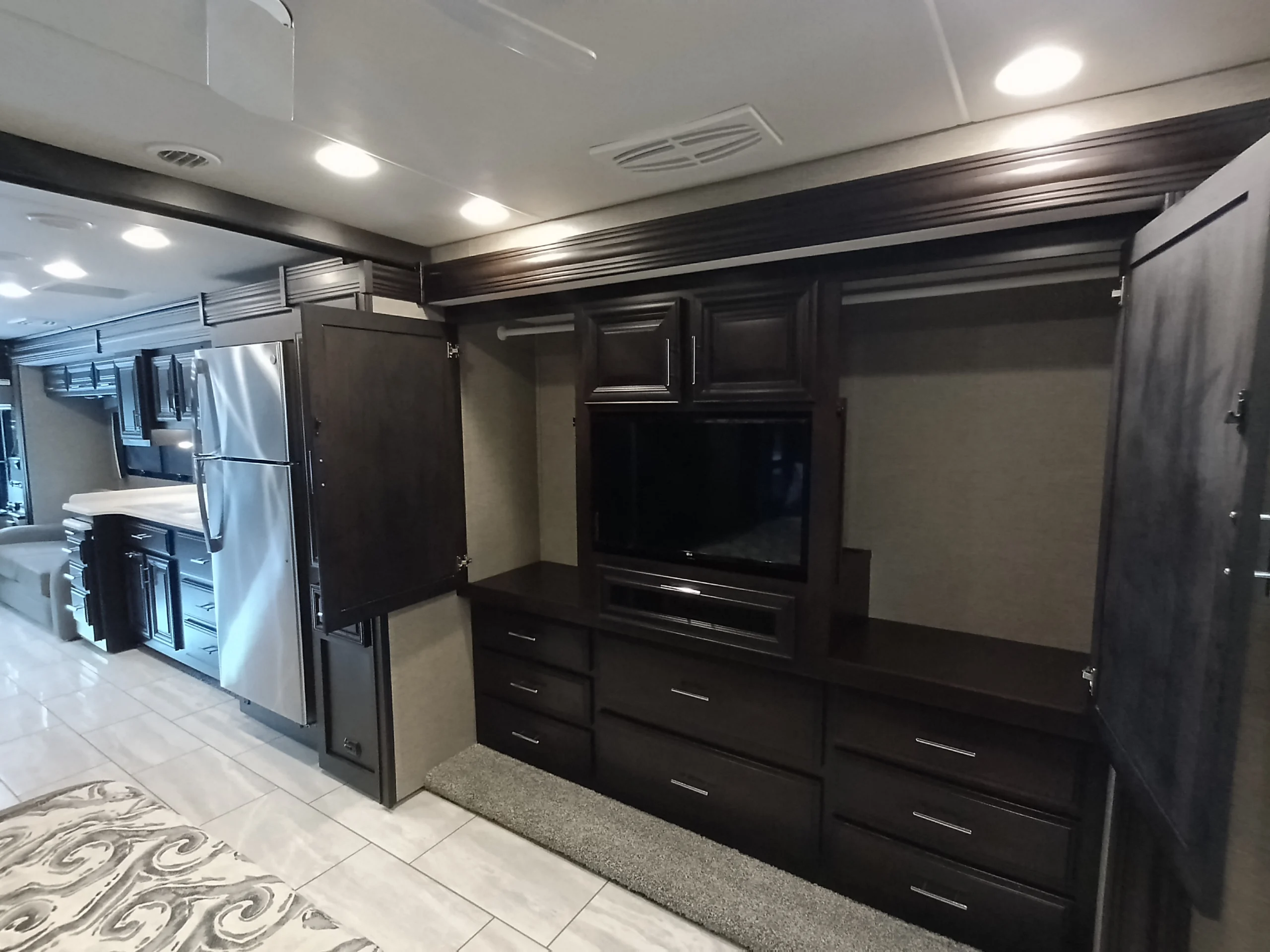 IMG_20260611_191816_159 Interior of a luxurious motorcoach featuring dark wood cabinetry, a stainless steel refrigerator, and ample storage space.
