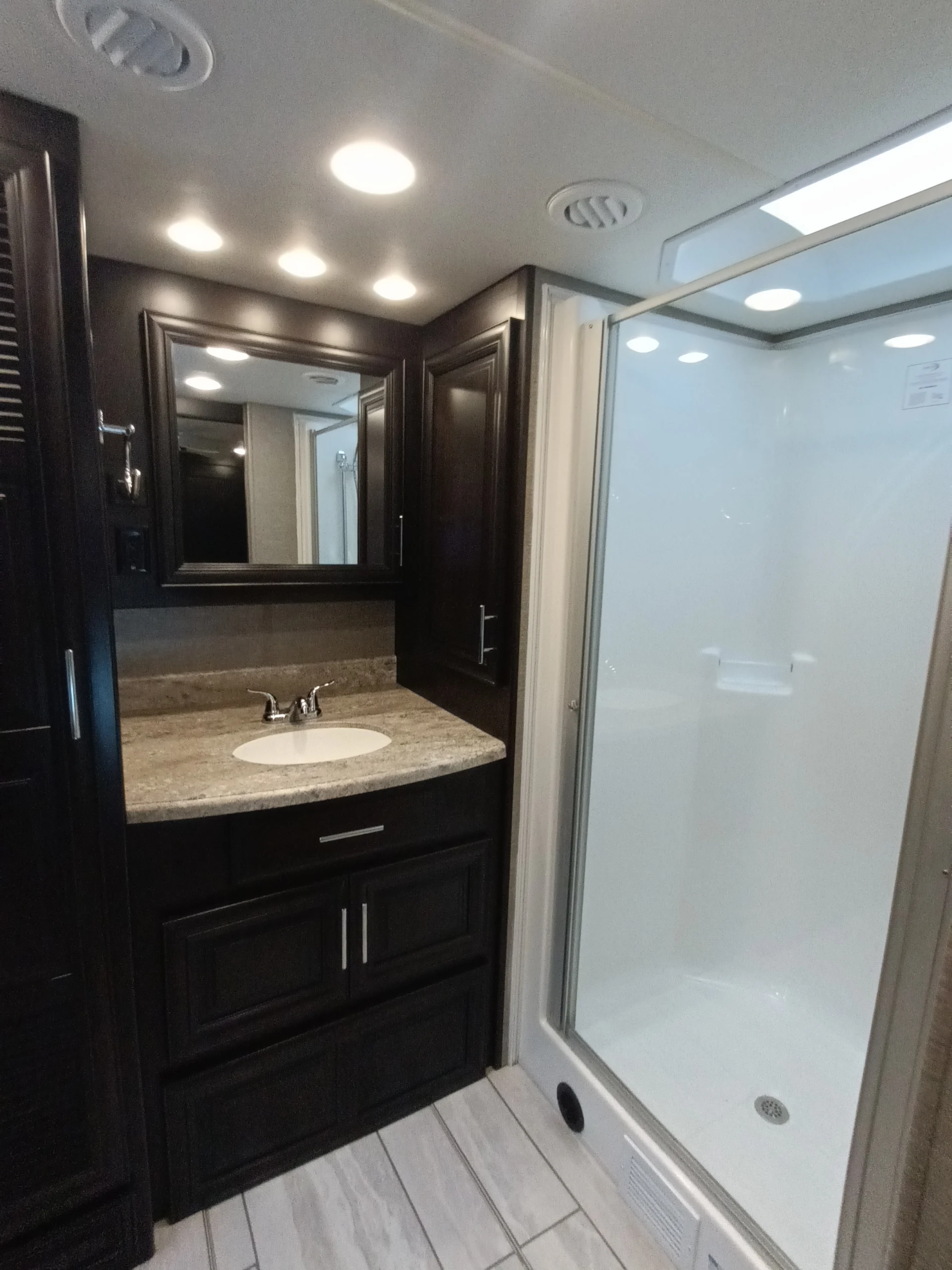 IMG_20260611_191908_654 RV bathroom with dark wood cabinets, a marble countertop sink, large mirror, and a glass-enclosed shower. Suitable for Motorcoach and RVs.