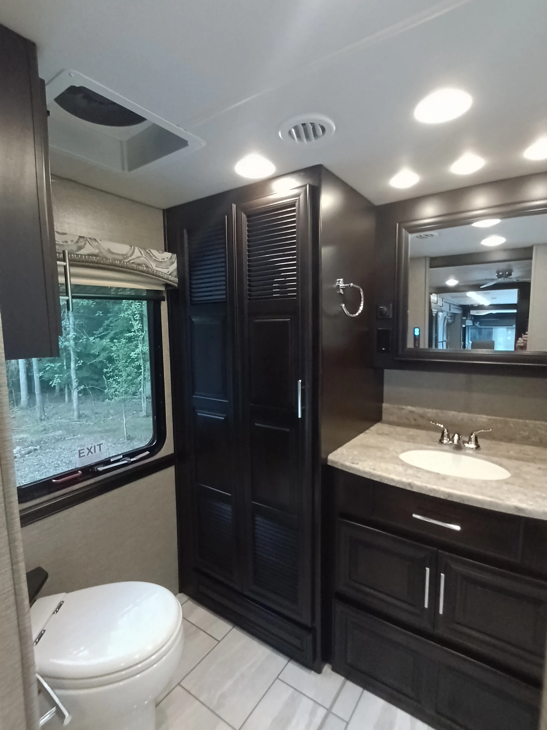 IMG_20260611_191920_596 Modern Motorcoach bathroom with dark wood cabinets, beige countertop, sink, toilet, and window with forest view. Recessed lighting above.