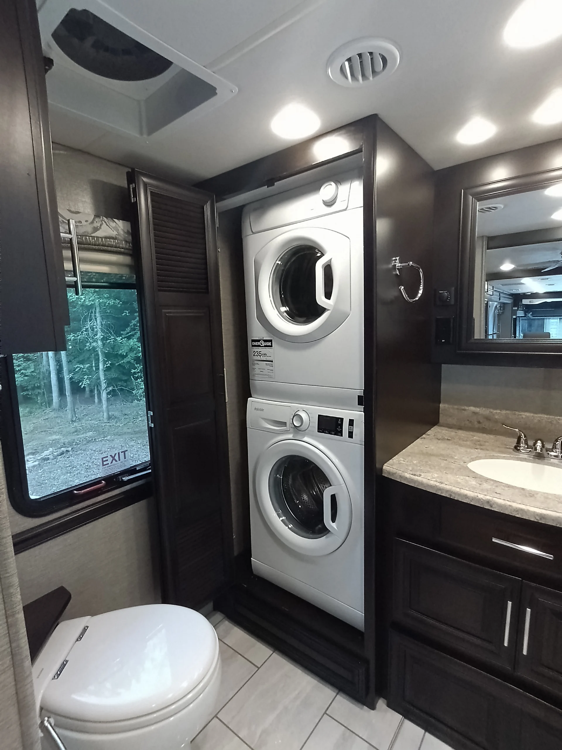IMG_20260611_191932_874 RV bathroom with stacked washer and dryer, sink, and toilet. Window with forest view, labeled exit. Ideal for Motorcoach and Rvs.