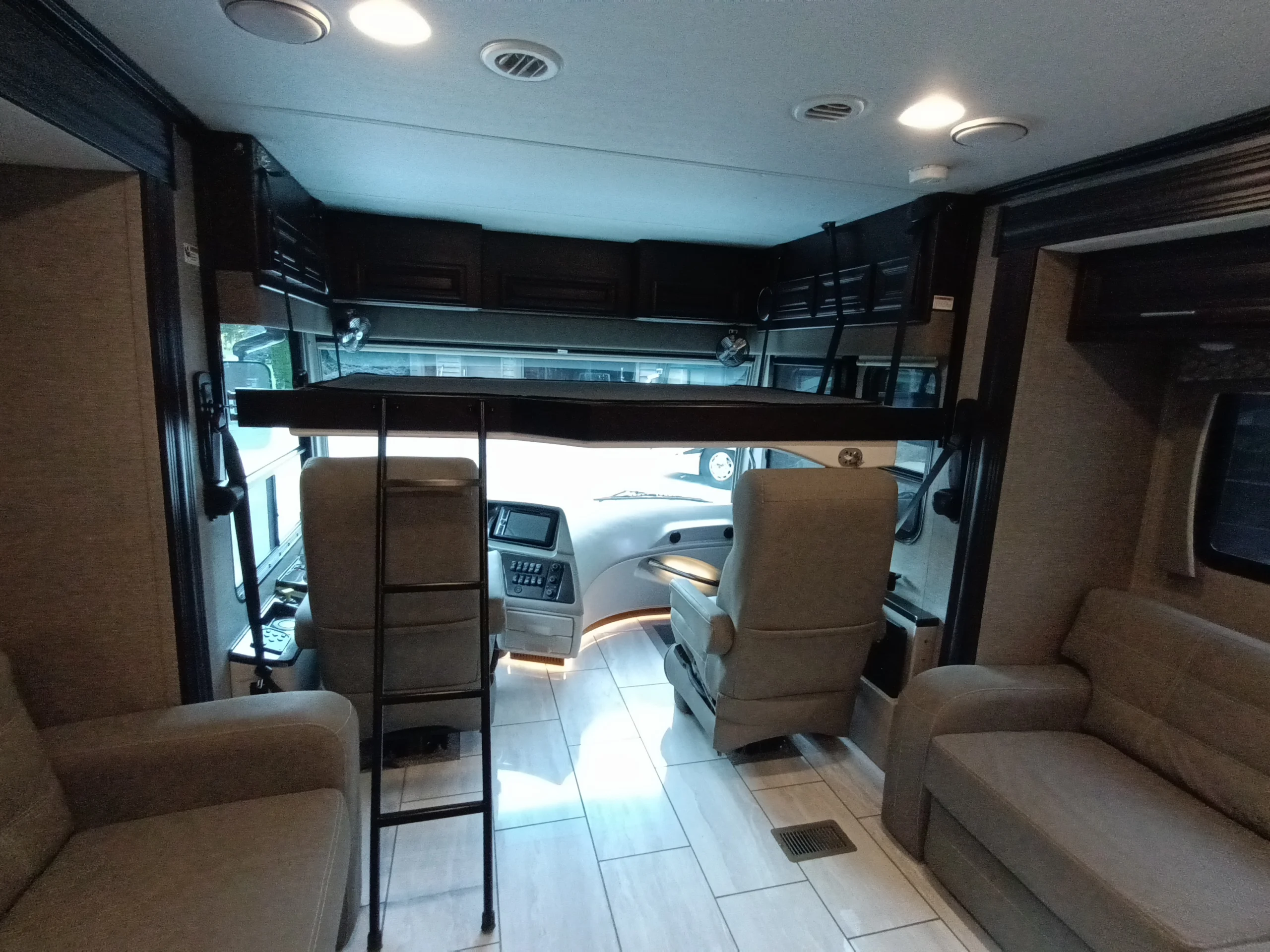 IMG_20260611_192216_848 Motorcoach interior with a driving area, two beige chairs, and a loft bed with ladder. Beige sofas line the walls.
