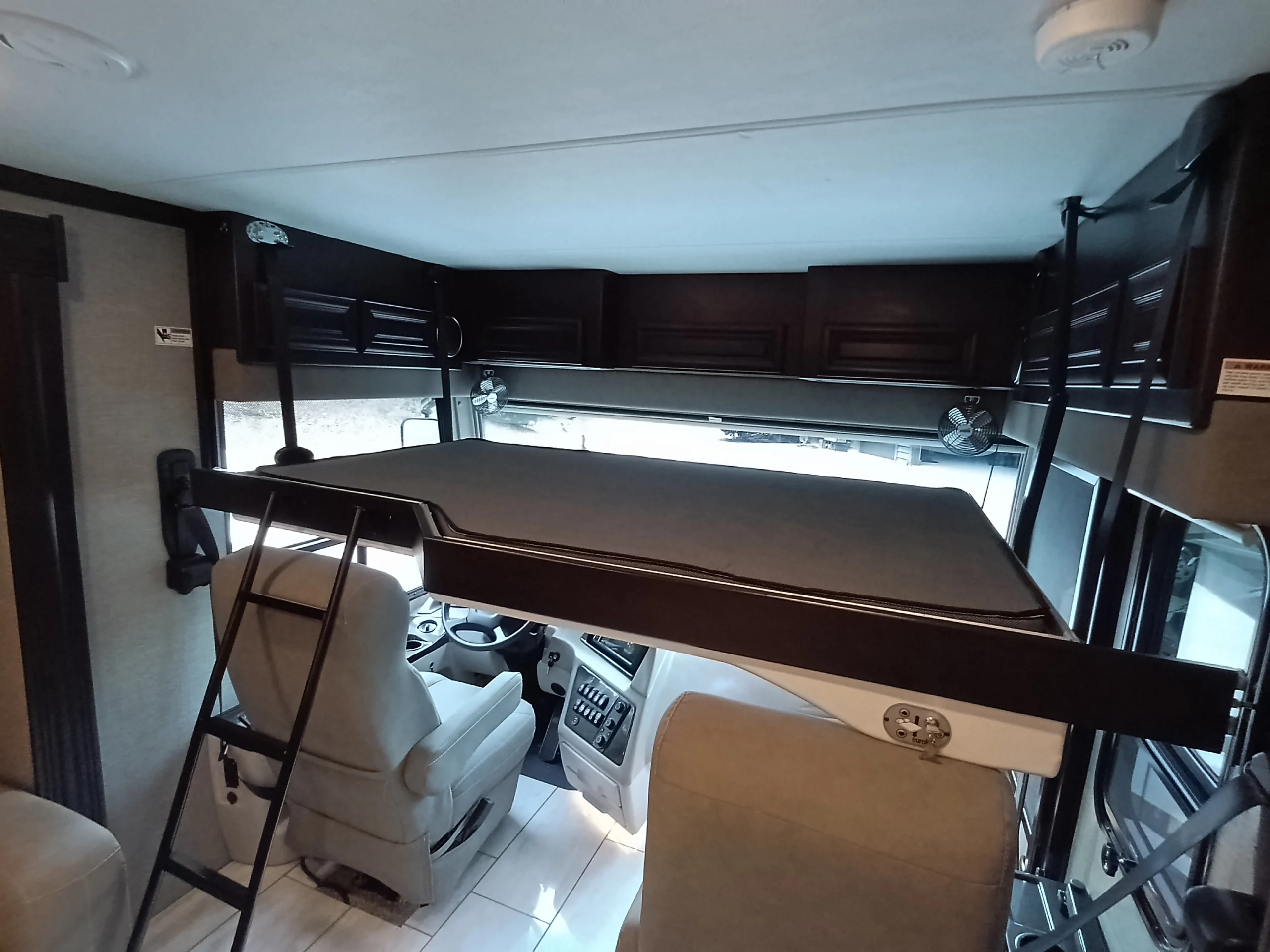 IMG_20260611_192224_421 Interior of a motorcoach featuring a loft bed with a ladder above the driver's seat, surrounded by dark wood cabinets.