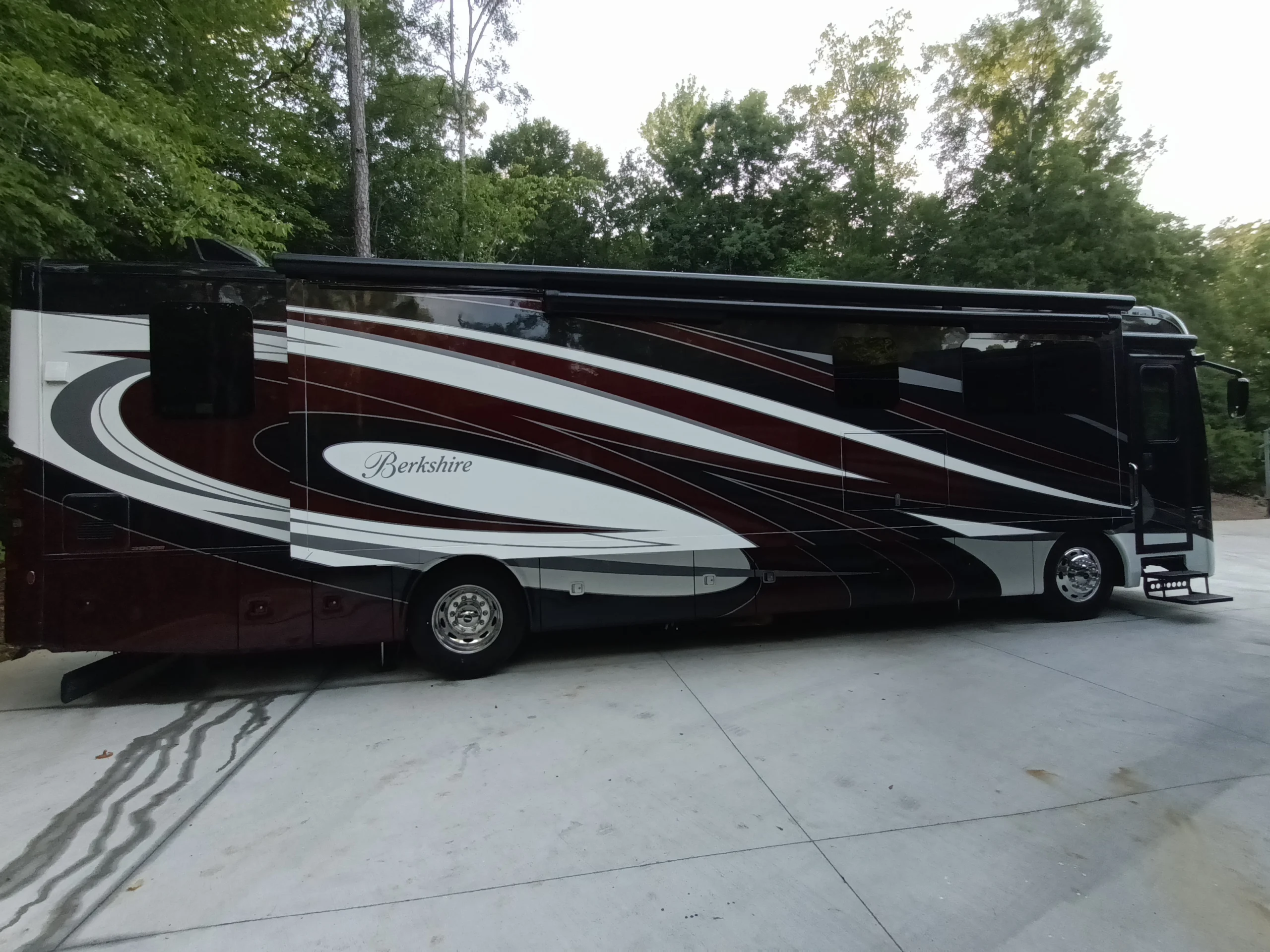 IMG_20260611_192531_256 Luxury Berkshire motorcoach parked on concrete, surrounded by trees, featuring sleek black, white, and maroon design.
