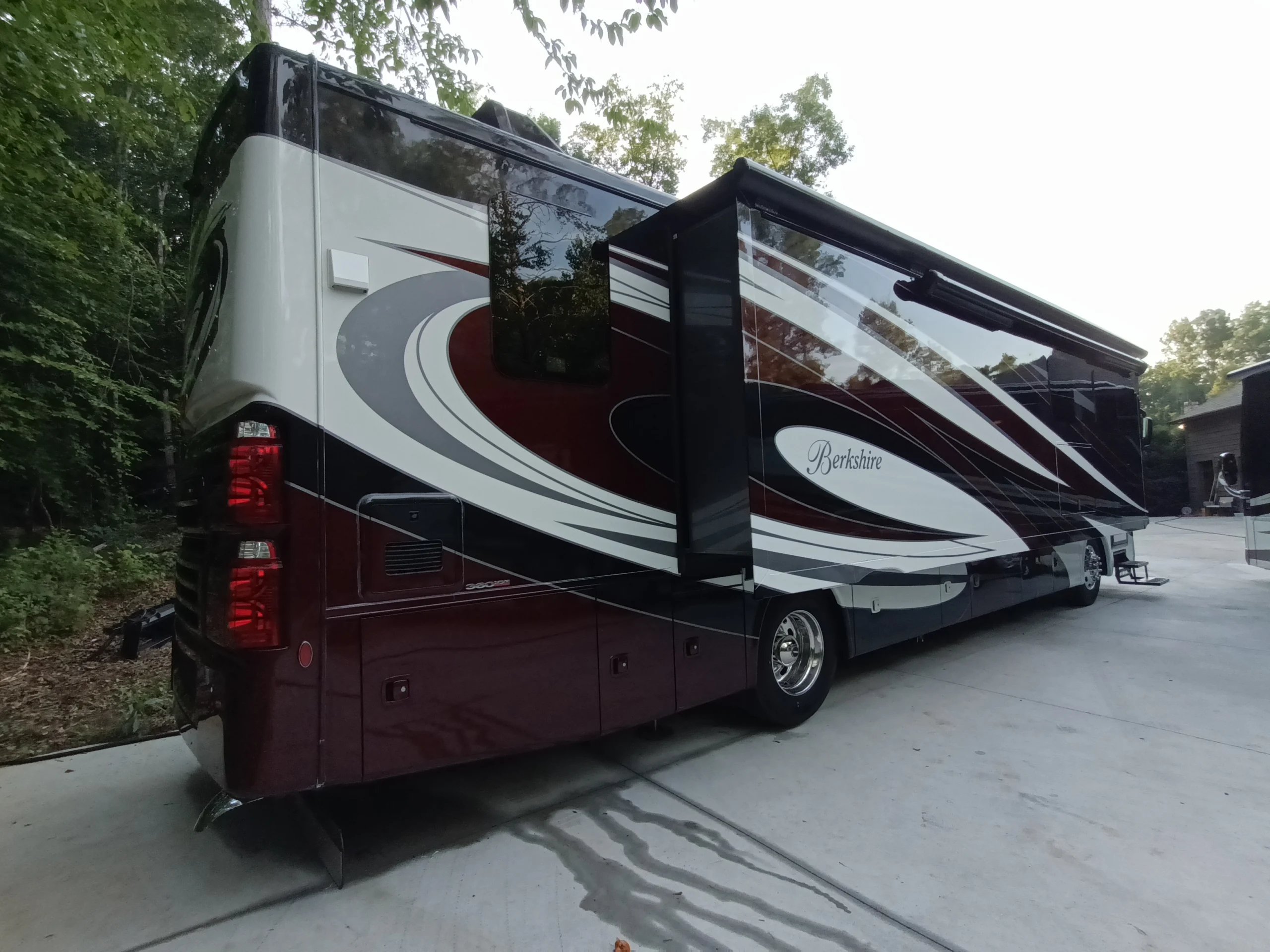 IMG_20260611_192541_750 Burgundy and white motorcoach parked on concrete with slide-out extended, surrounded by trees and a building.