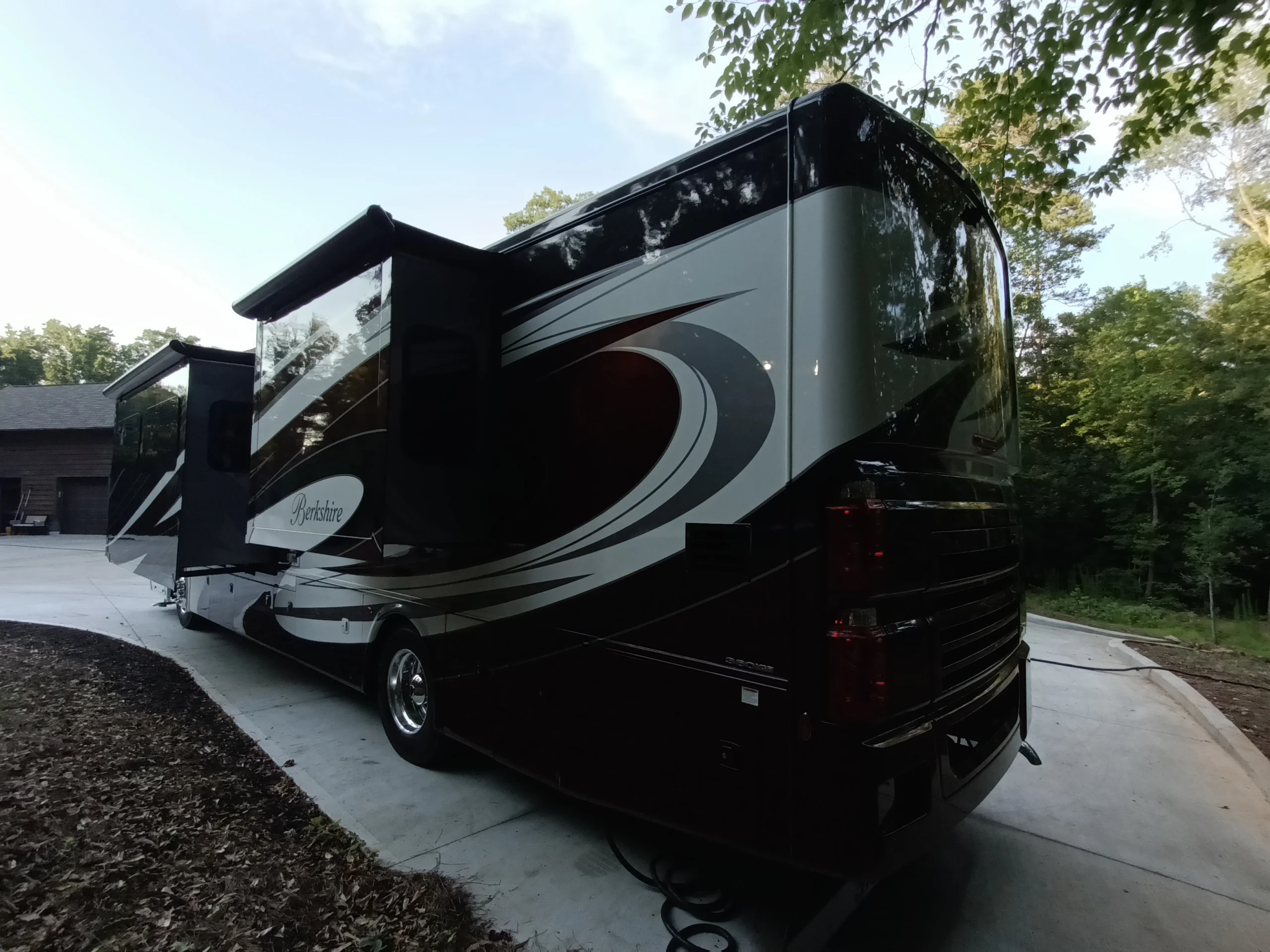 IMG_20260611_192555_580 Luxury Motorcoach parked on a concrete driveway near a garage, surrounded by trees under a clear sky.