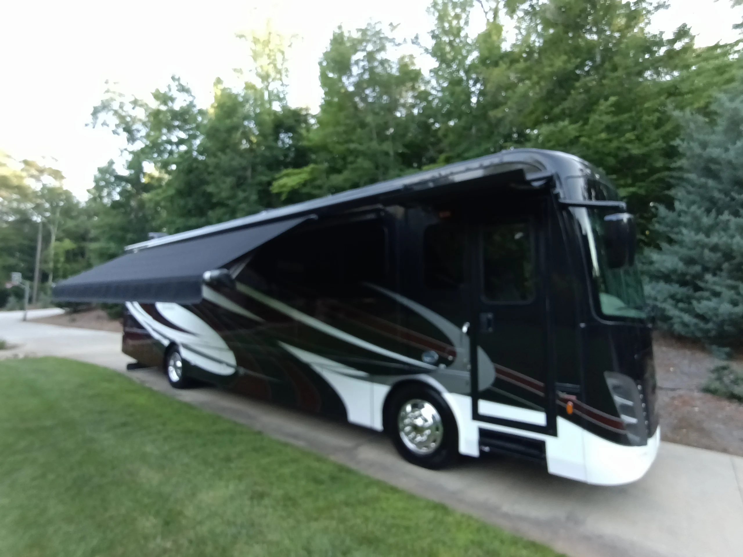 IMG_20260611_193818_948 Black motorcoach with an extended awning, parked on a driveway, surrounded by lush green trees.