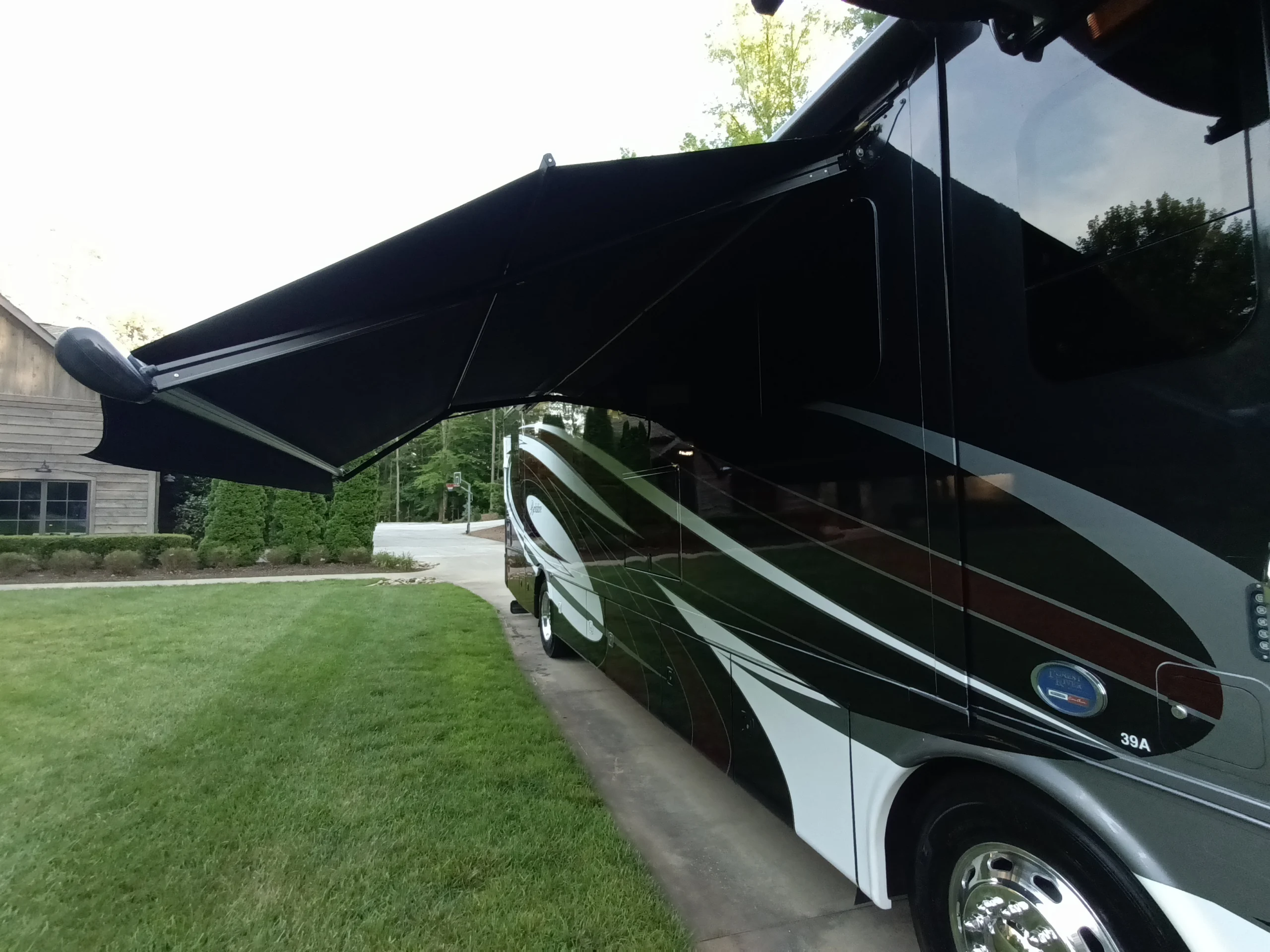 IMG_20260611_193825_744 Motorcoach with extended black awning parked on driveway, adjacent to a neatly mowed lawn and wooden building.