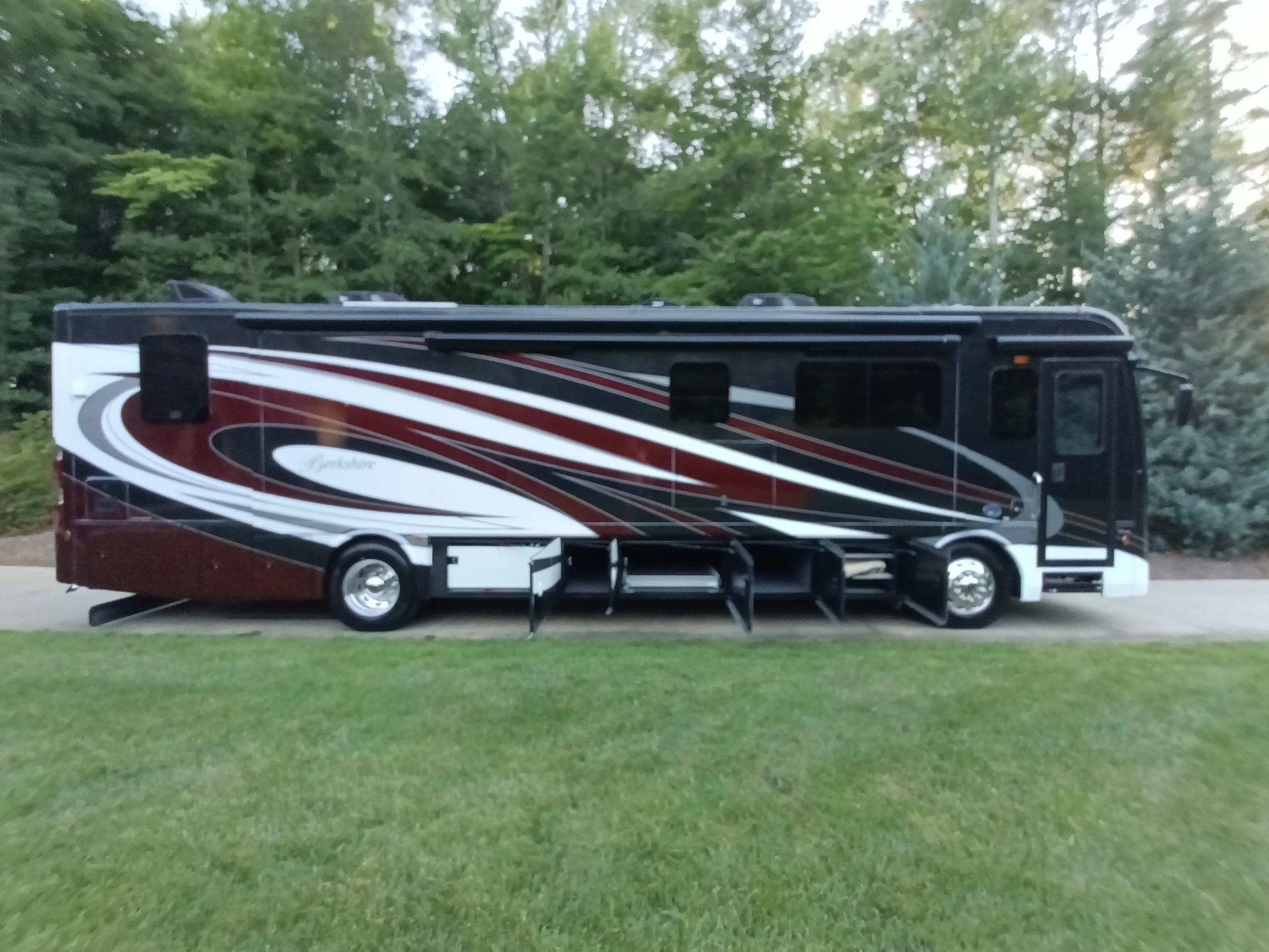 IMG_20260611_193954_876 Luxurious motorcoach with maroon, black, and white swirls, parked on grass with trees in the background.