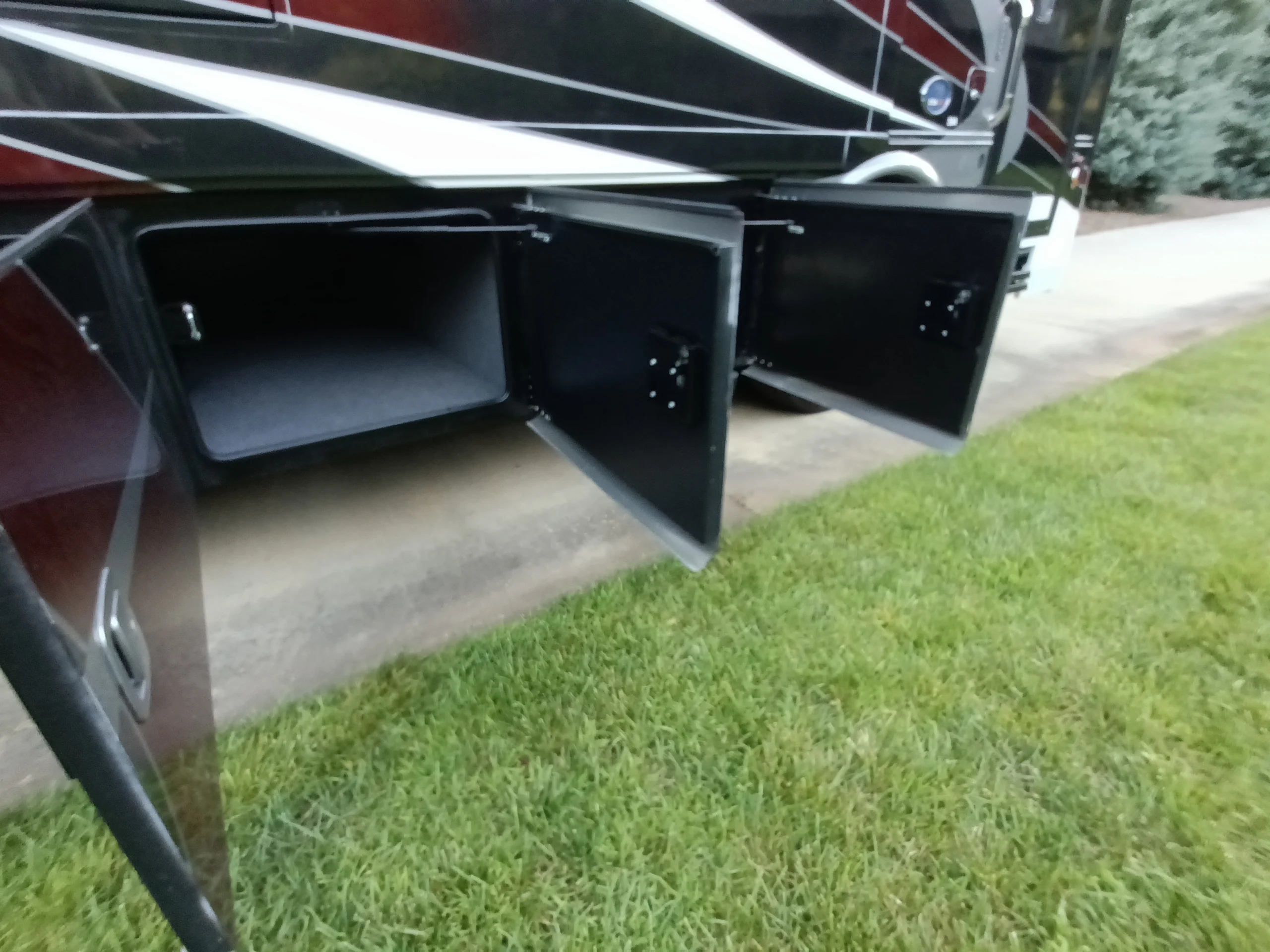 IMG_20260611_194003_733 Open storage compartments under a Motorcoach and RVs, revealing ample space for gear. Nearby, grass and pavement present.
