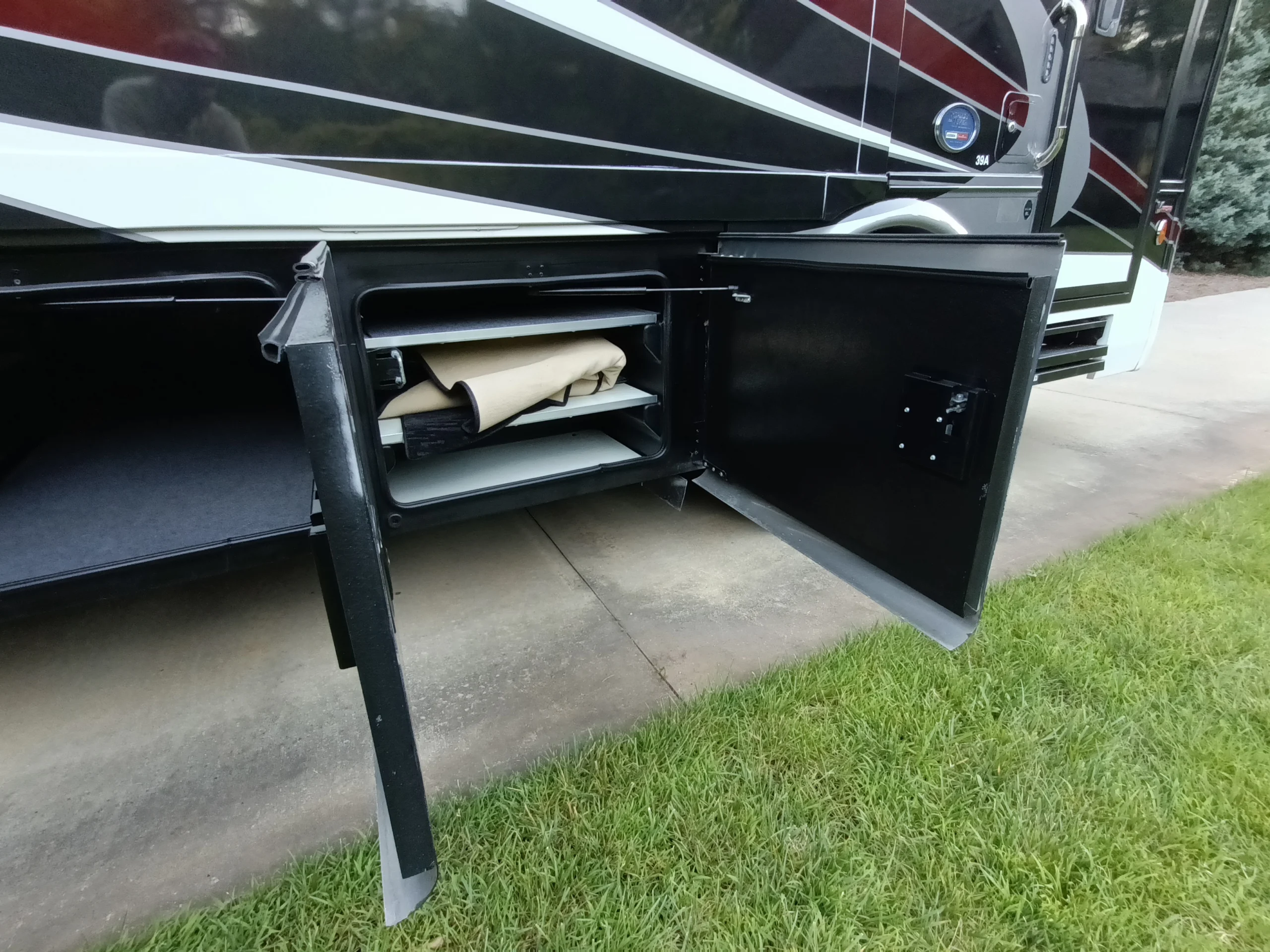 IMG_20260611_194008_970 Open storage compartment in a Motorcoach and RVs showing shelves with folded item, parked on a driveway beside grass.