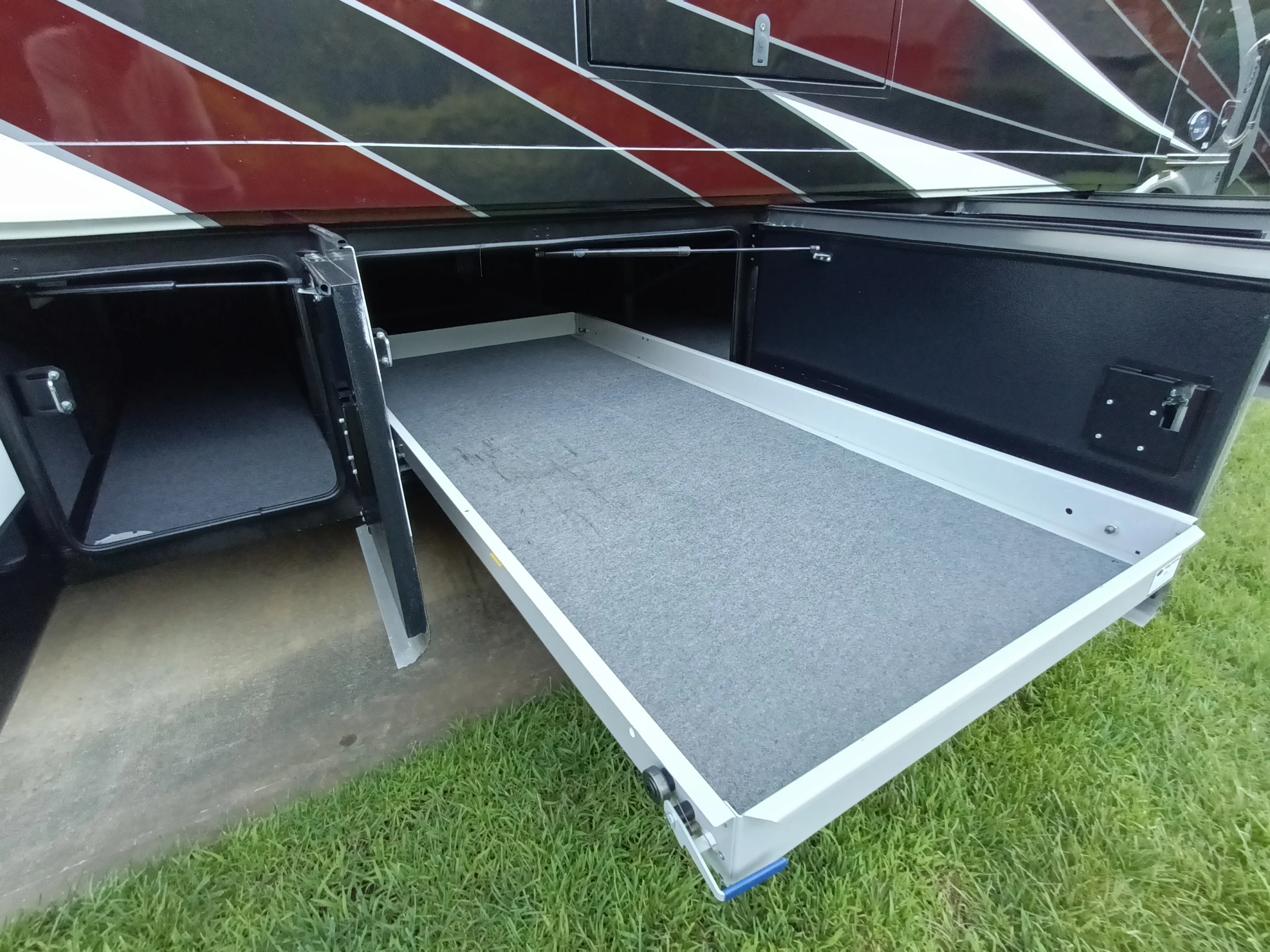 IMG_20260611_194019_767 Open storage compartment with slide-out tray on a Motorcoach and RVs unit, parked on grass.