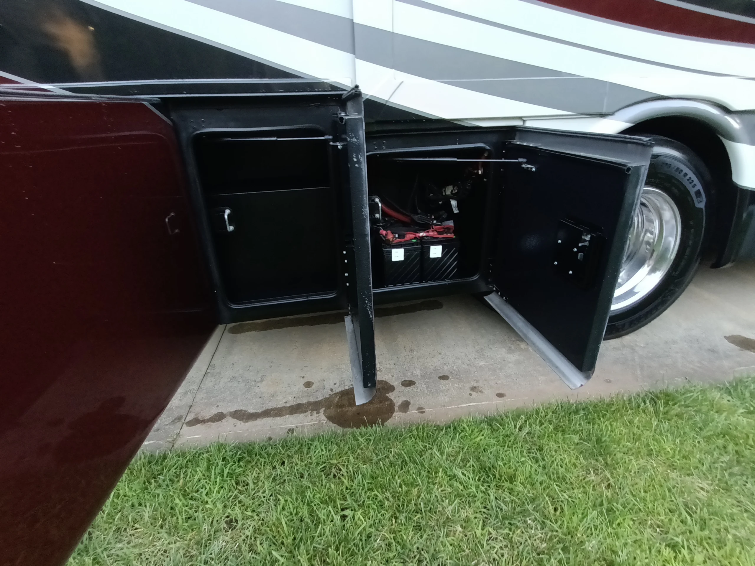 IMG_20260611_194110_048 Open storage compartments near the wheel of a Motorcoach, displaying batteries inside, with grass and concrete surrounding.