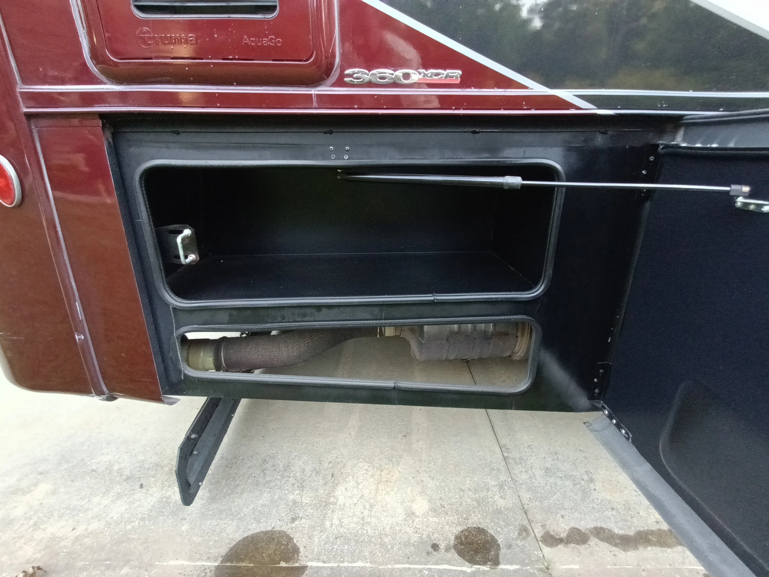 IMG_20260611_194119_698 Open storage compartment on a motorcoach, revealing internal shelving and a pipe below, exterior painted in maroon with a logo.