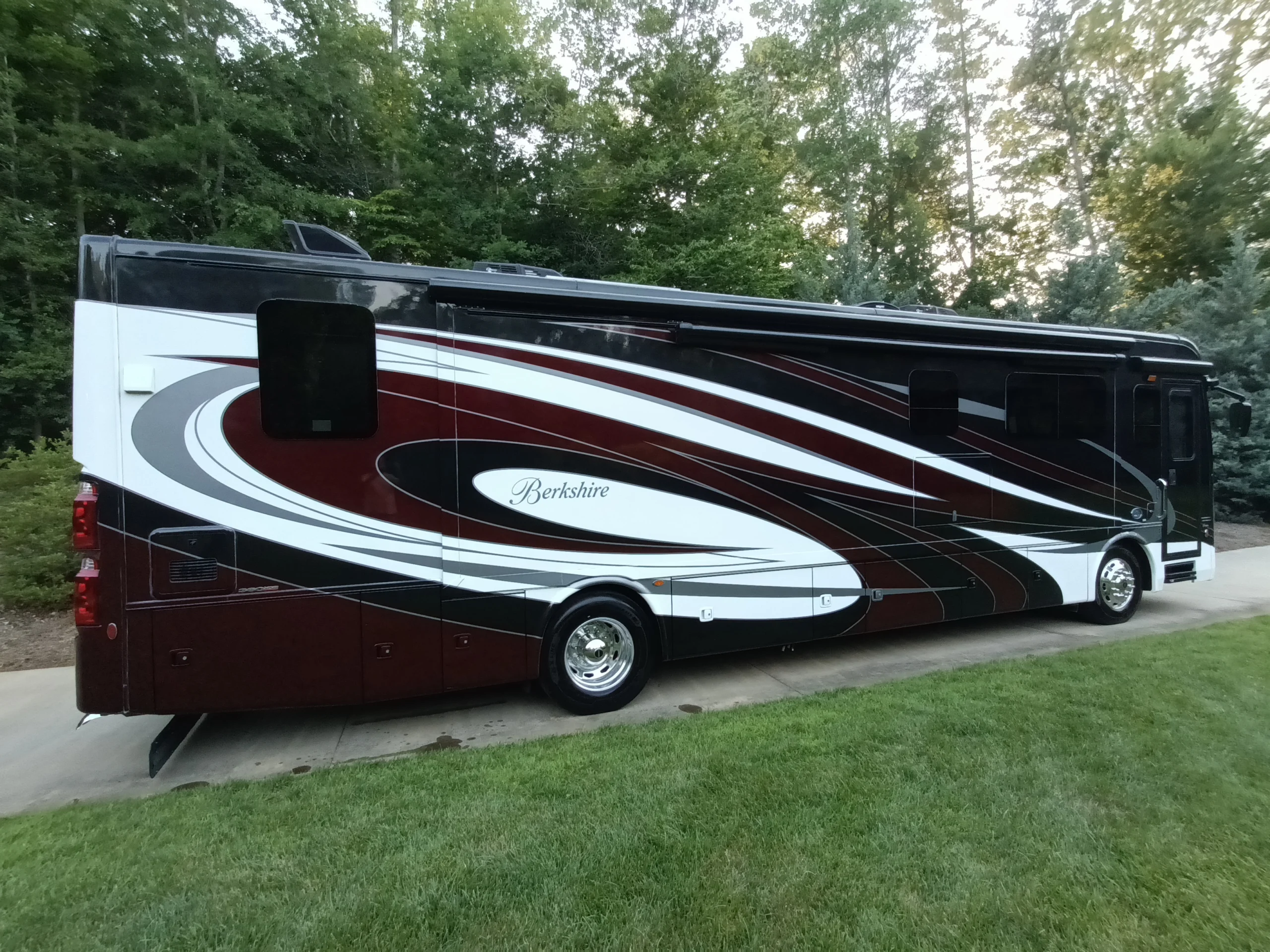 IMG_20260611_194222_285 Luxurious motorcoach with sleek red, black, and white design parked on a driveway, surrounded by lush green trees and grass.
