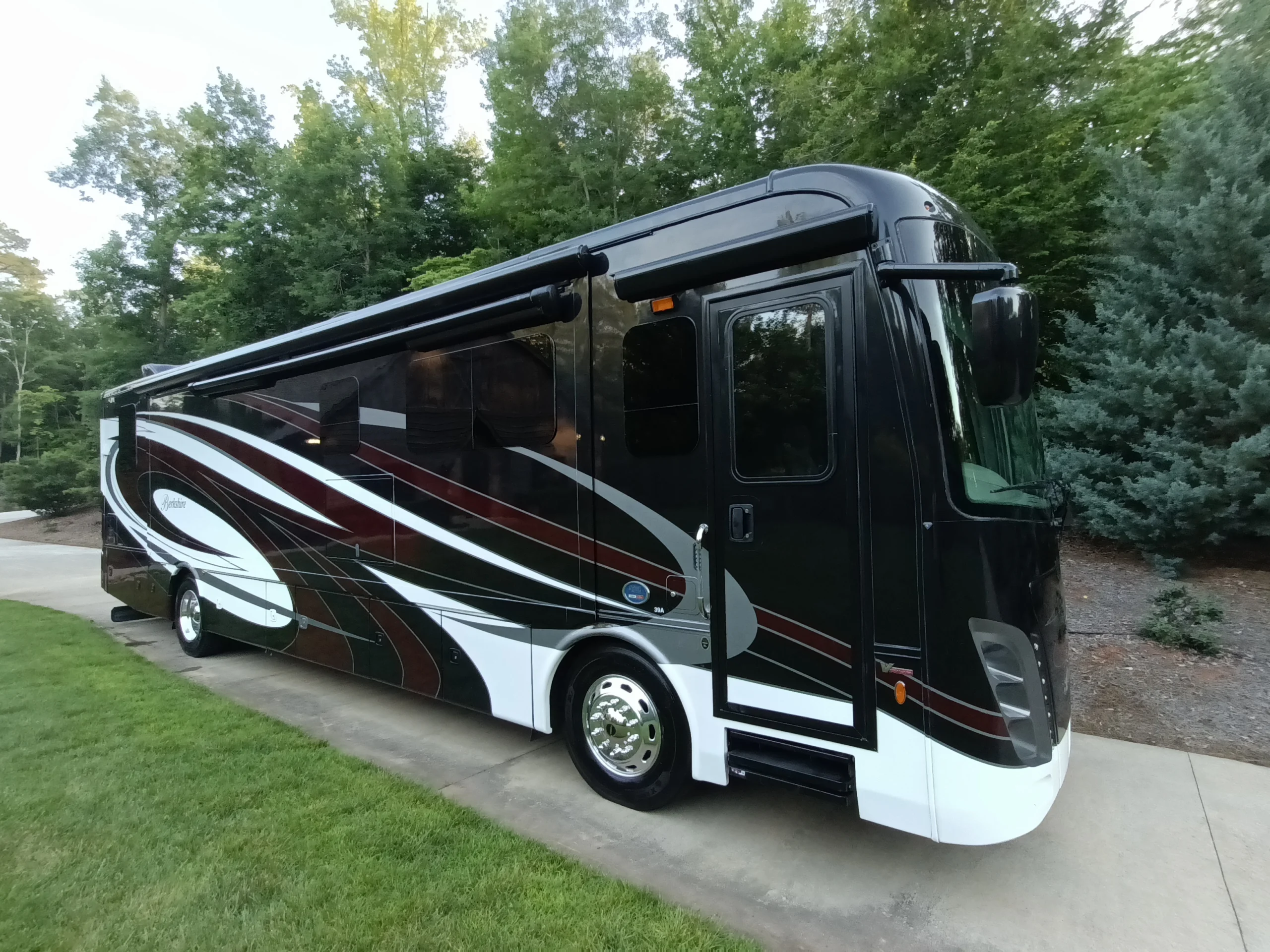 IMG_20260611_194244_633 Sleek black Motorcoach and RV with bold stripes parked on a driveway, surrounded by lush green trees and grass.