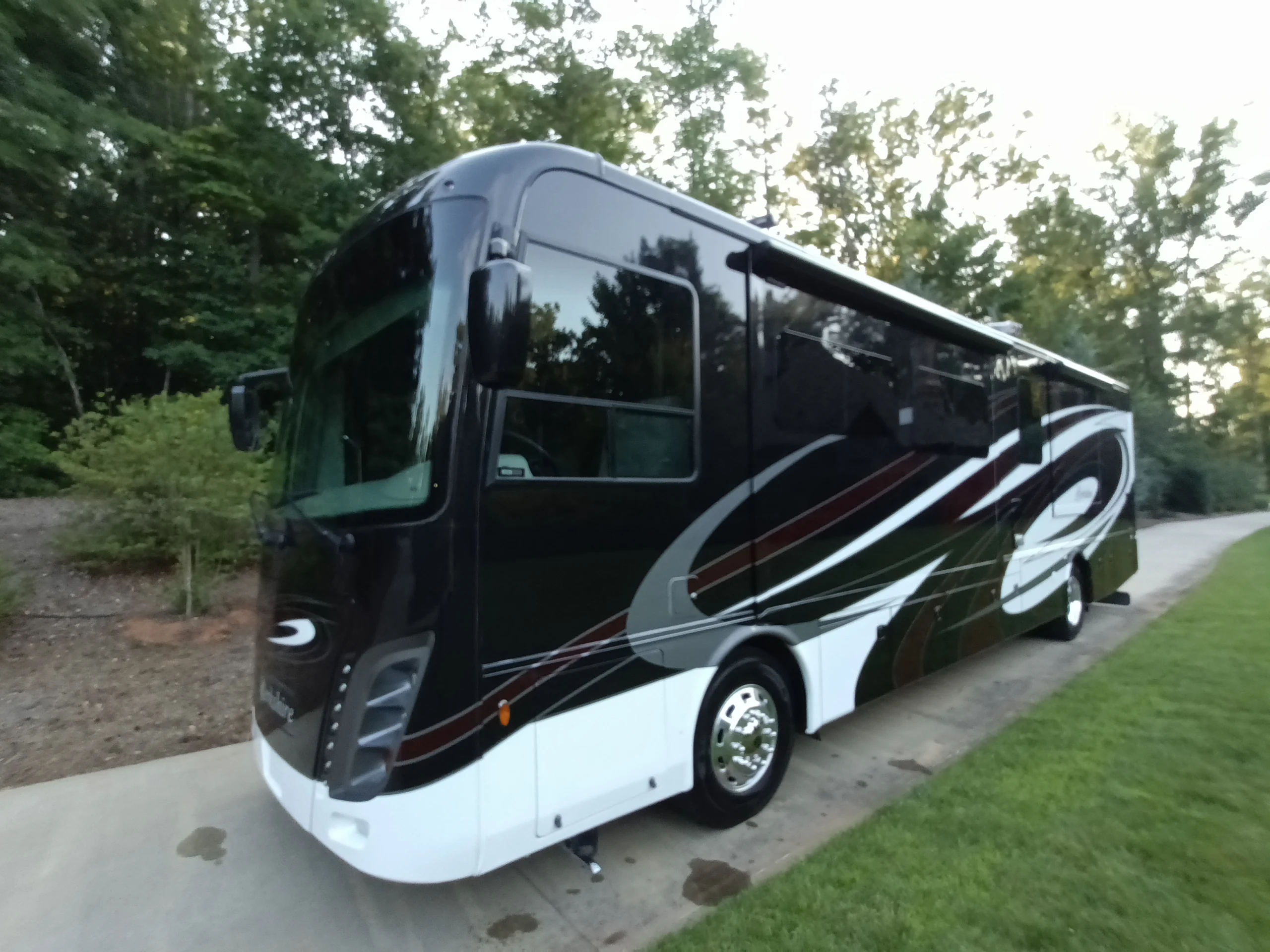 IMG_20260611_194640_071 Sleek black motorcoach RV with modern graphics parked on concrete, surrounded by lush greenery.