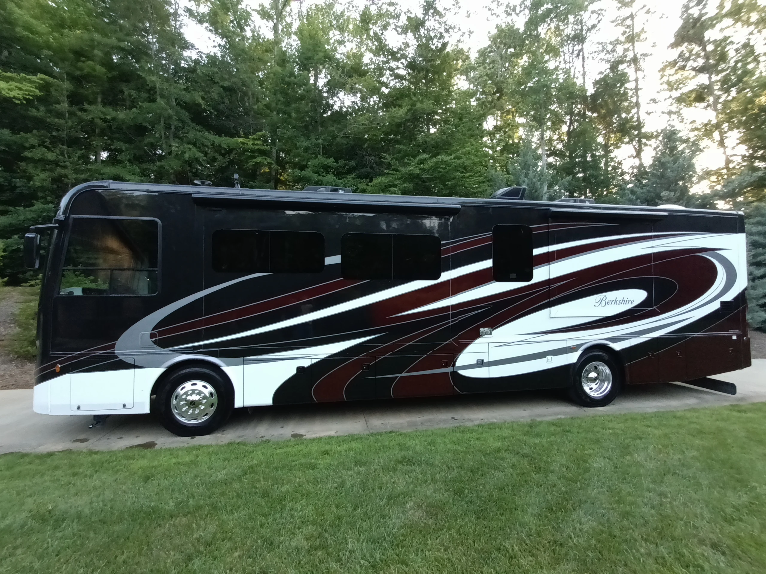 IMG_20260611_194650_214 Sleek black and white motorcoach with dynamic red and white swirls parked on a driveway, surrounded by lush greenery.