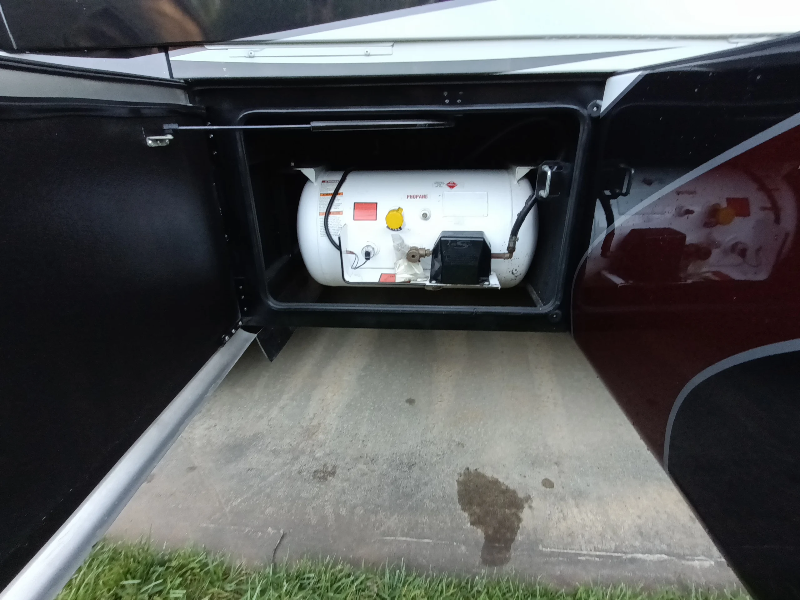 IMG_20260611_194754_415 Open compartment in a motorcoach revealing a propane tank with gauges and connections, situated under a concrete surface.