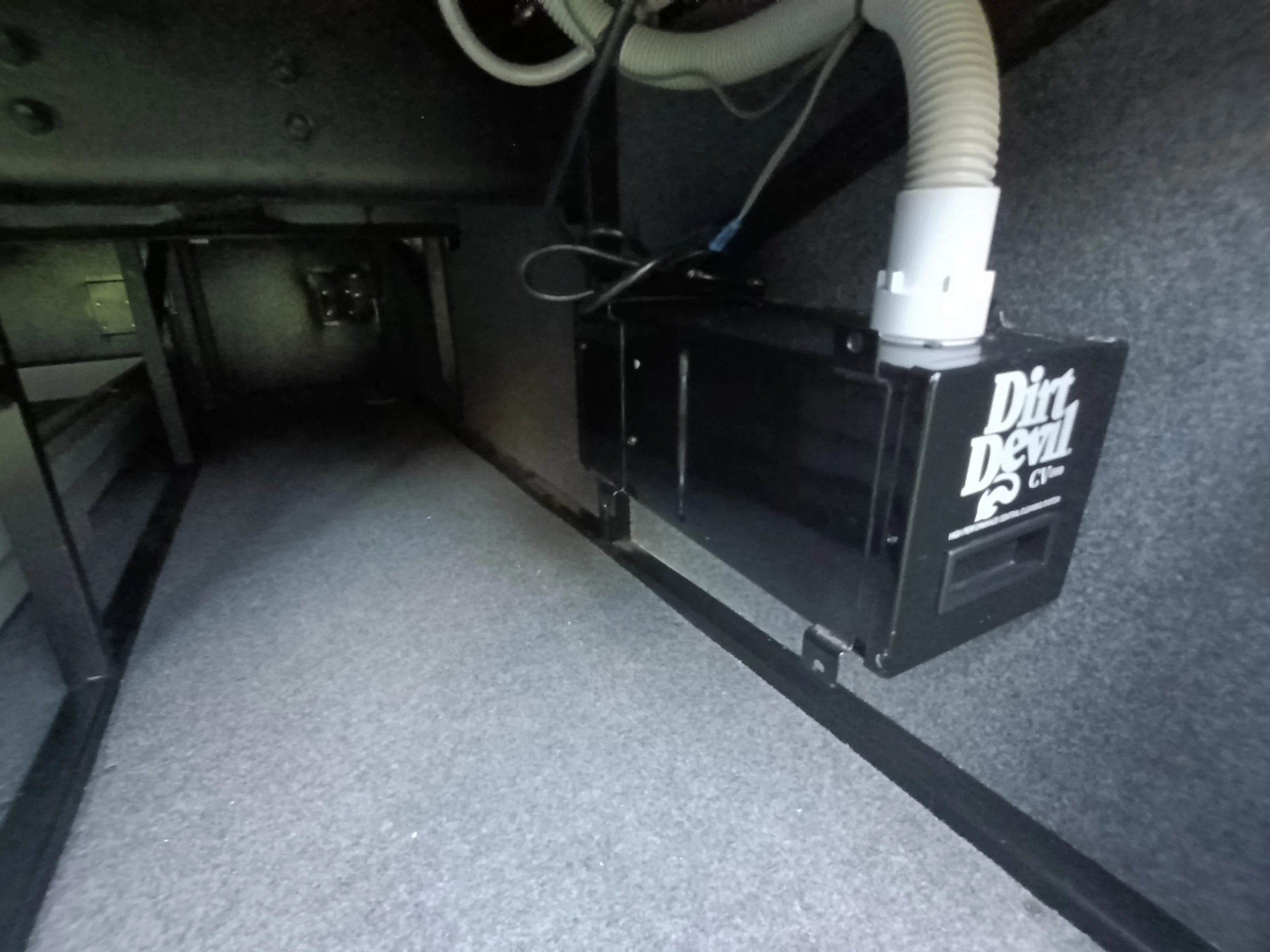IMG_20260611_194827_211 Central vacuum system labeled "Dirt Devil" installed in RV storage compartment. Grey carpet lining the walls and floor.