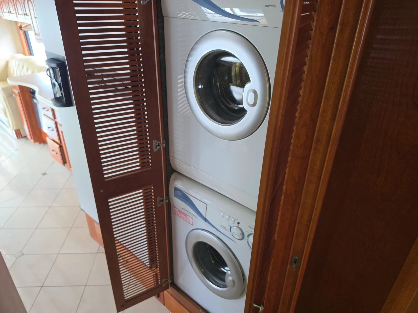 20260831171445-adc413ea-7741-4e42-bd70-49724ec283a2 Stacked washer and dryer in a Motorcoach and Rvs, behind wooden slatted doors, in an interior with beige tile and cabinetry.