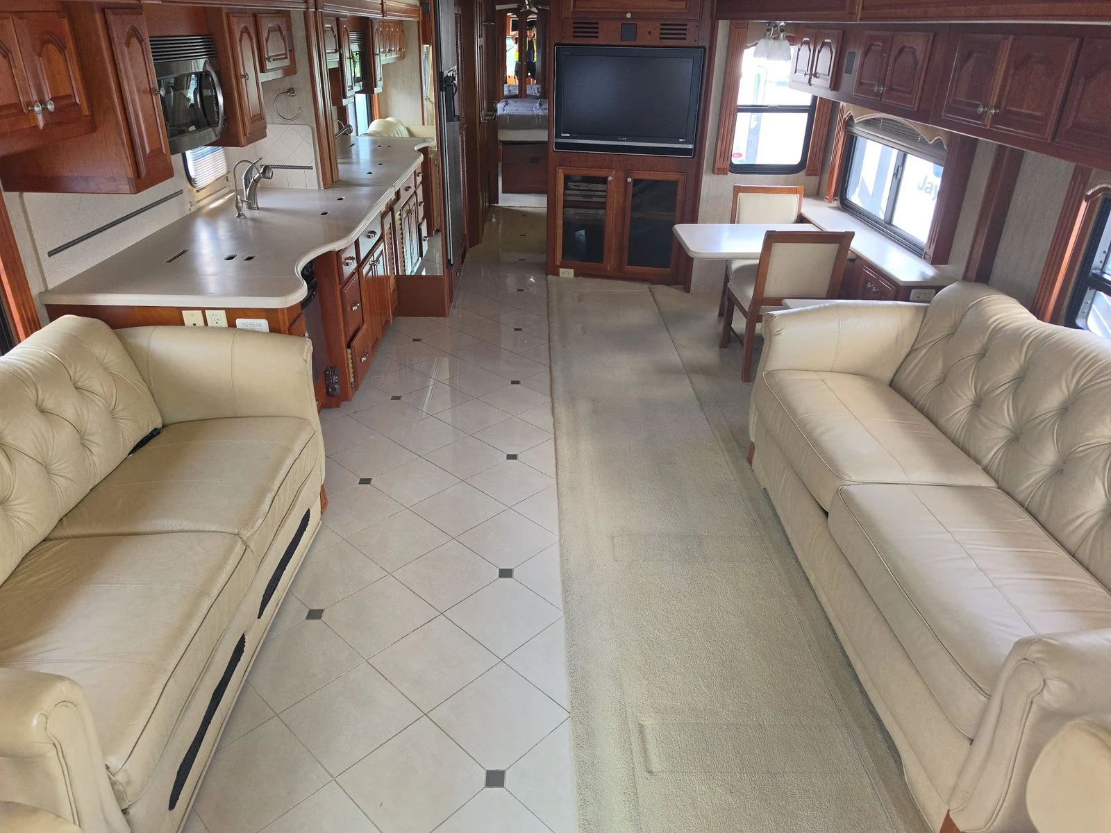 20260831171446-00650afb-d861-4da8-b26b-23c119c8abd1 Spacious Motorcoach interior with cream leather sofas, tiled floor, wood cabinetry, kitchen area, dining table, and wall-mounted TV.