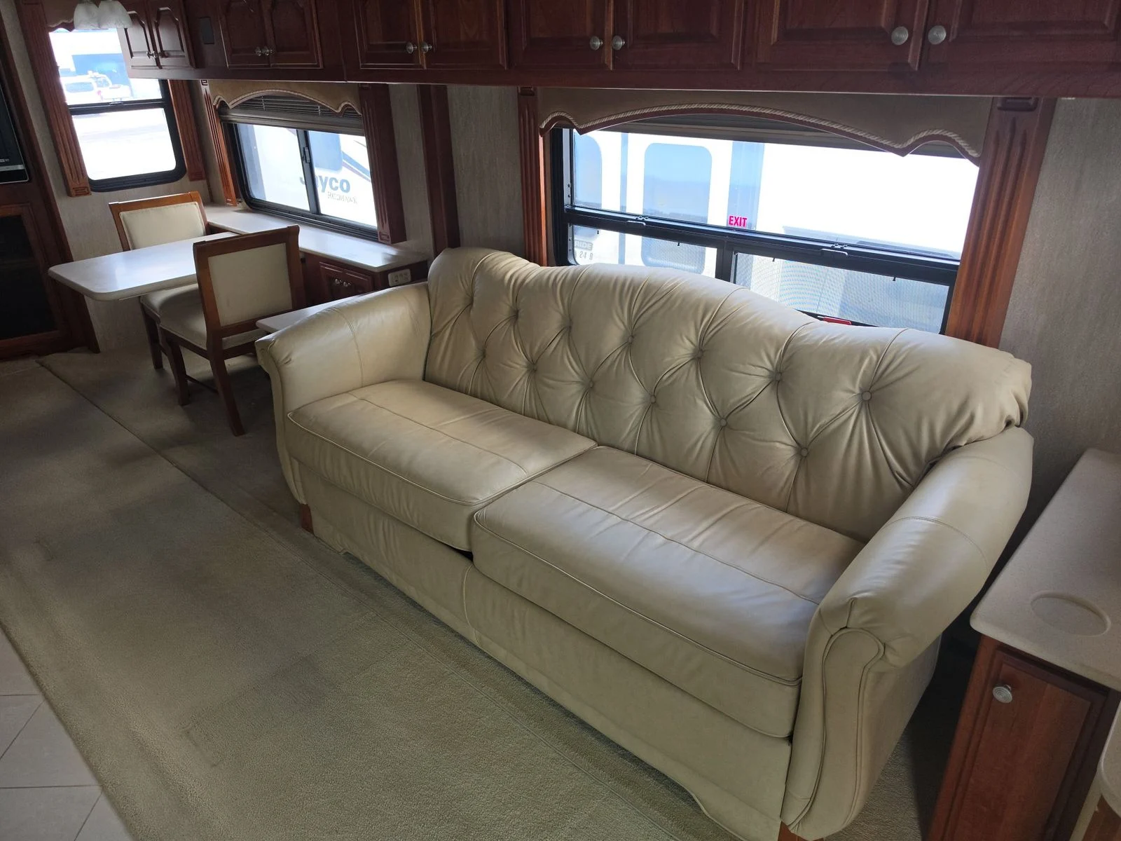 20260831171446-5d57bb95-5cb2-456b-bc05-31f5572fbfea Luxurious Motorcoach and RVs interior with a plush cream leather sofa, dining table, and chairs, surrounded by wooden cabinetry and large windows.