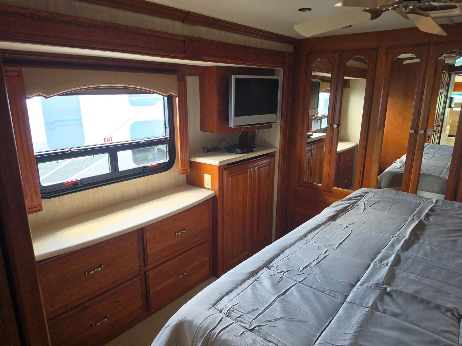 20260831171450-6ea2d885-3a63-4139-8dbd-705d3e41c313 Spacious Motorcoach bedroom with wooden cabinetry, a TV, mirrored closet doors, and a window. Rv exterior visible outside.