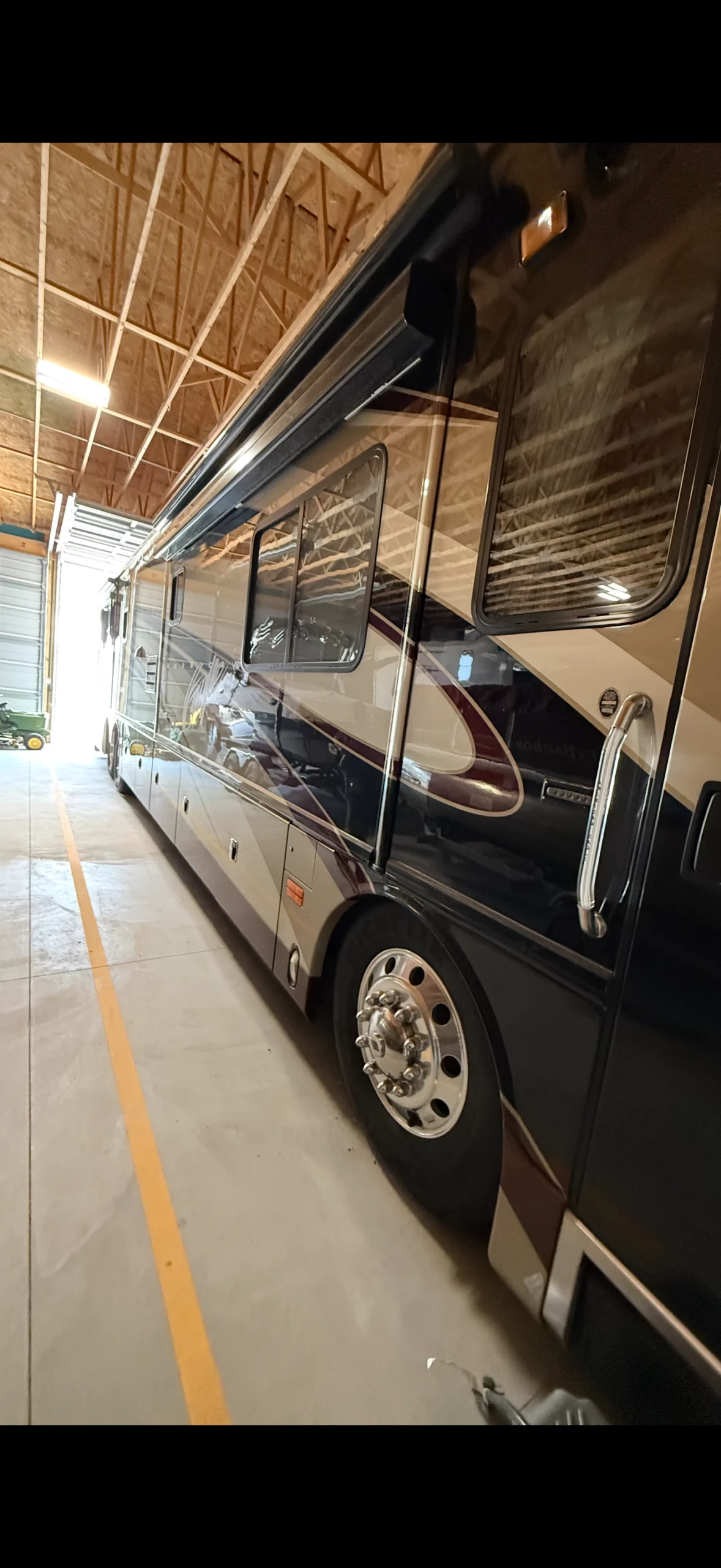 IMG_20260905_114425 Large motorcoach parked in a spacious garage, showing sleek side profile with windows, under high wooden ceiling and soft lighting.