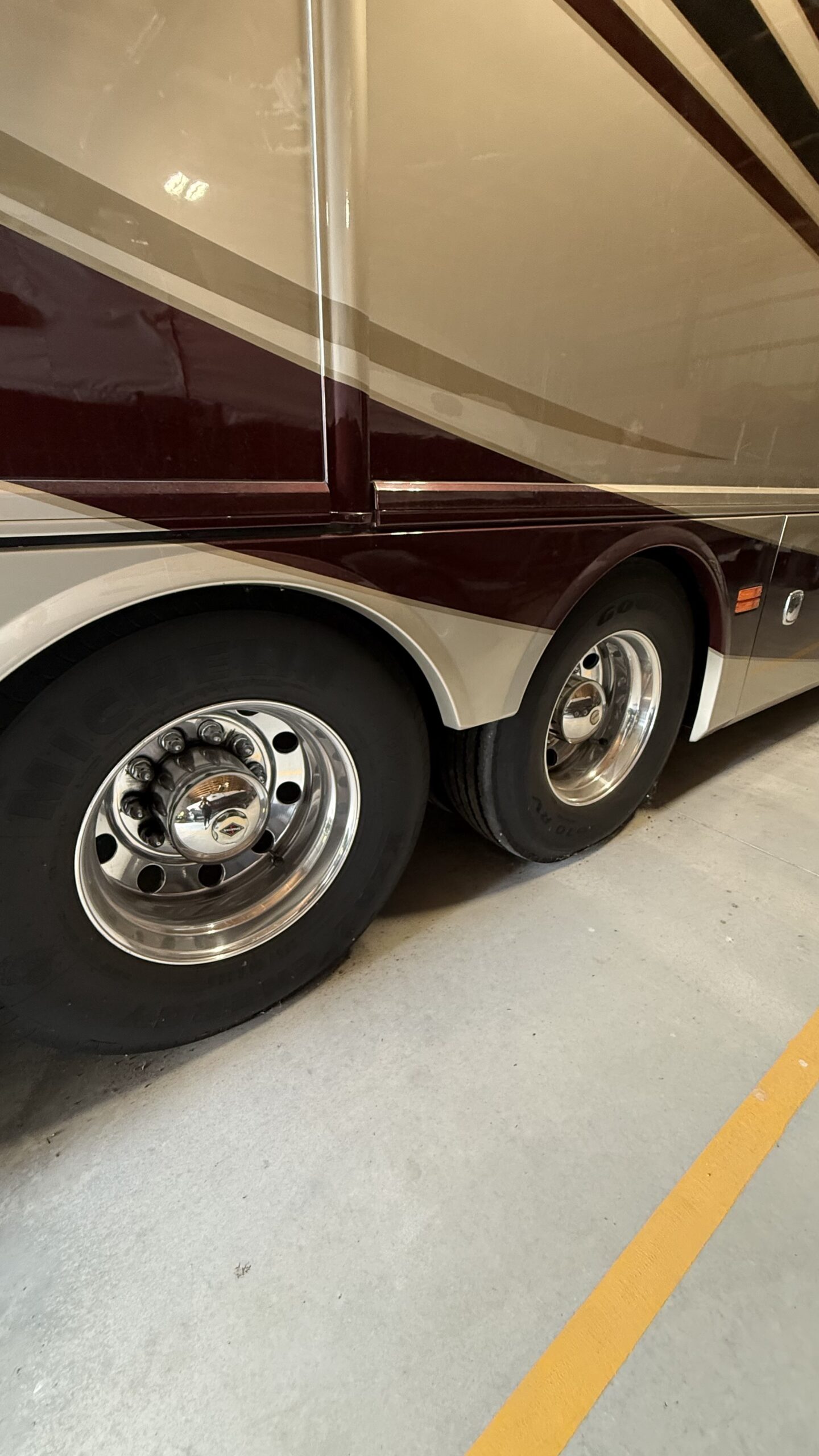 IMG_20260905_114450 Close-up of a motorcoach's rear wheels and tire details, parked indoors on a concrete floor with a yellow line.