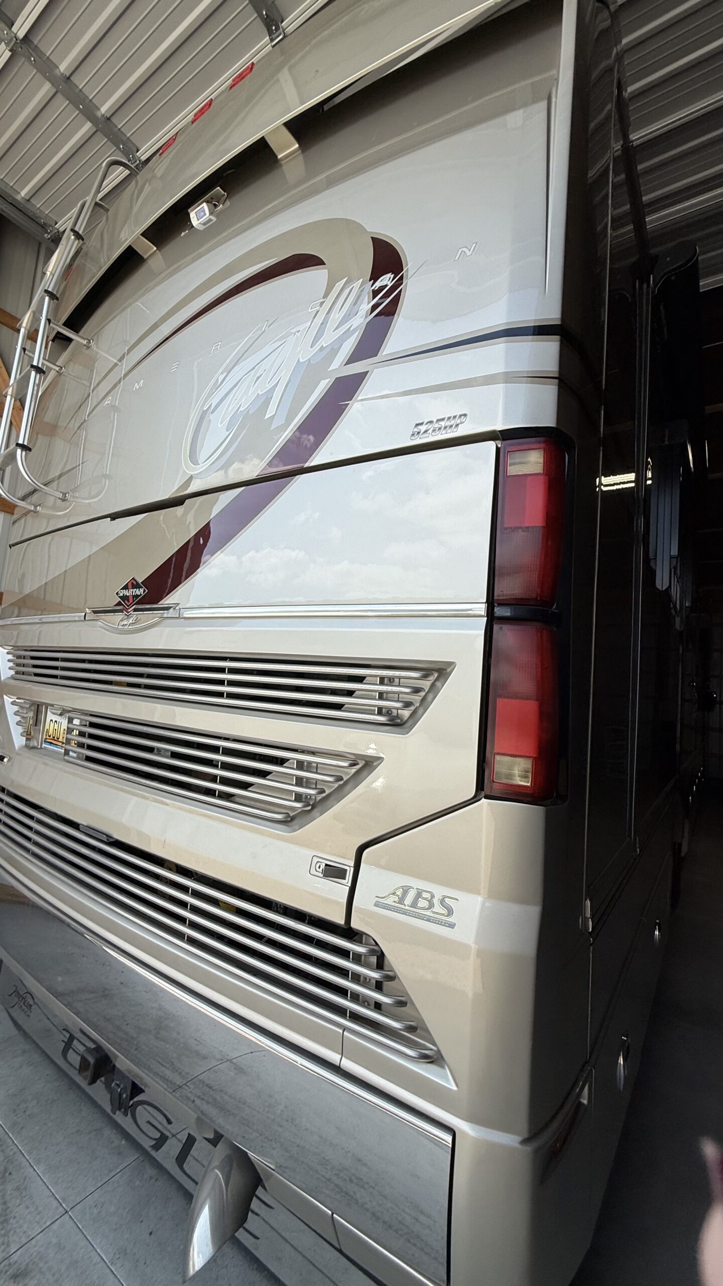 IMG_20260905_114501 Rear view of a parked luxury motorcoach with a ladder, aerodynamic design, and bold red accents, inside a garage.