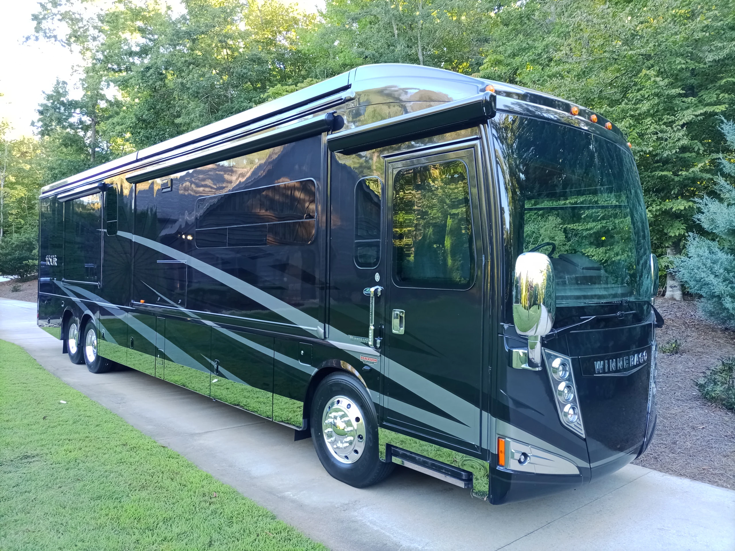 IMG_20260928_171921_579 Sleek black motorcoach with silver accents parked on concrete driveway, surrounded by lush green trees.