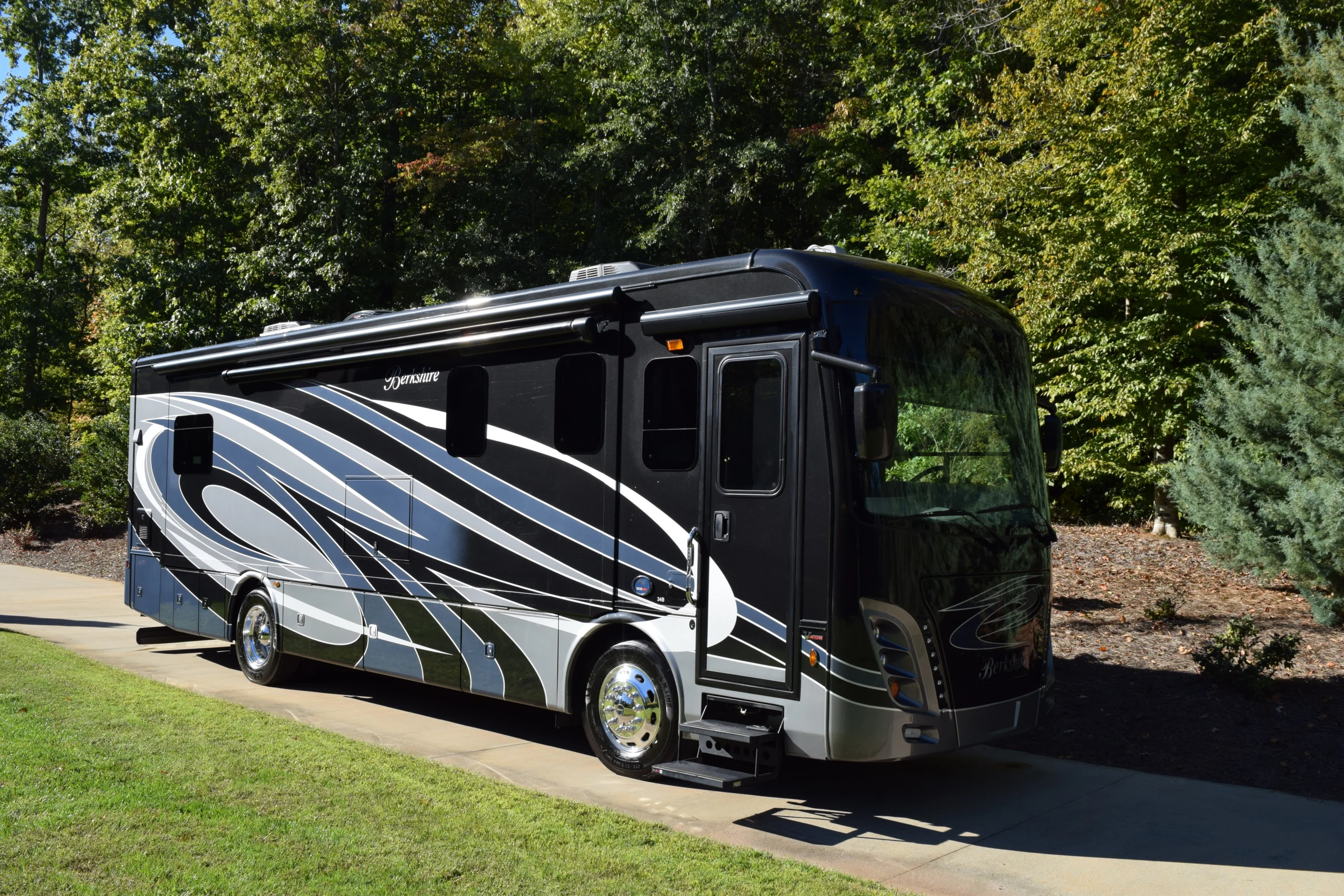 Luxurious motorcoach parked on a sunlit, paved driveway, surrounded by lush green trees and grass.