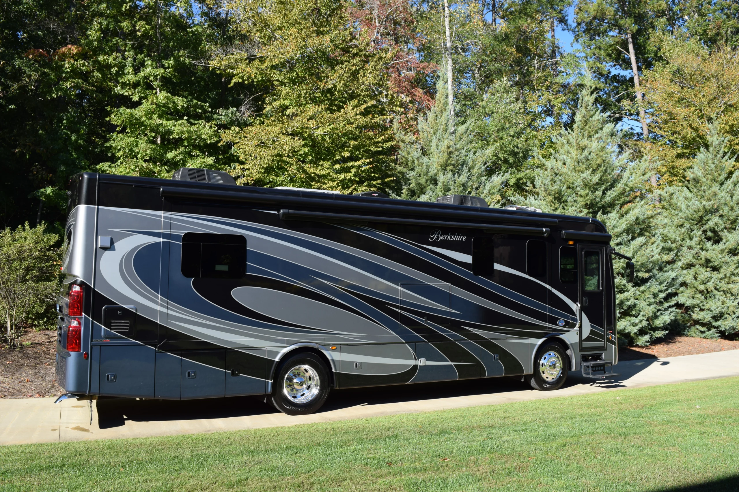 DSC_0691 Motorcoach with sleek silver and gray design parked on a driveway, surrounded by lush green trees.